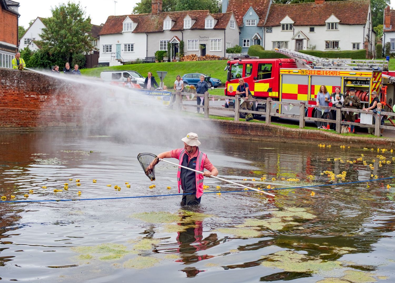 Person wading in flooded village pond collecting rubber ducks while fire engine sprays water during charity duck race event.