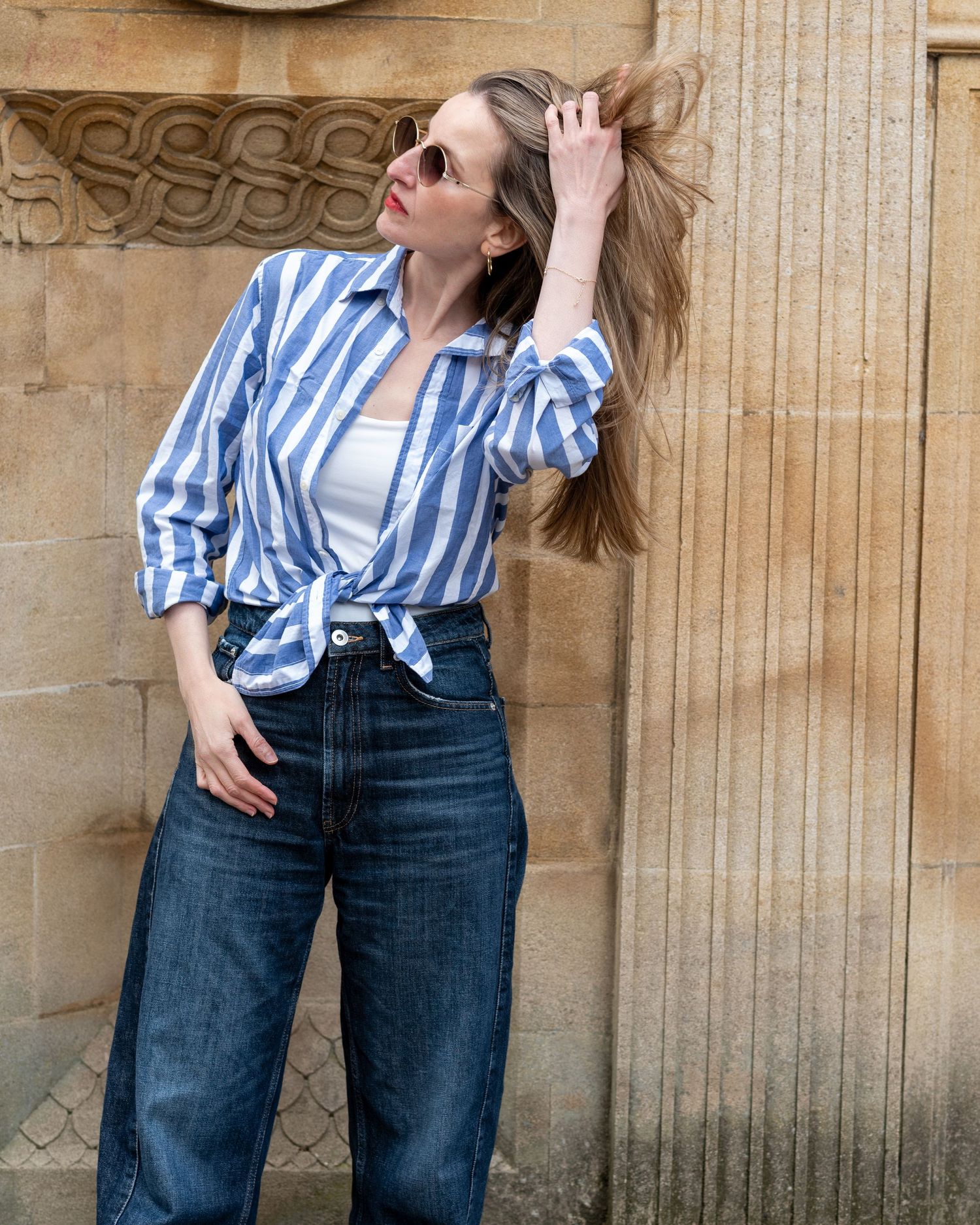 Woman in blue striped shirt tied at waist, white top, and dark jeans posing against stone wall wearing sunglasses.
