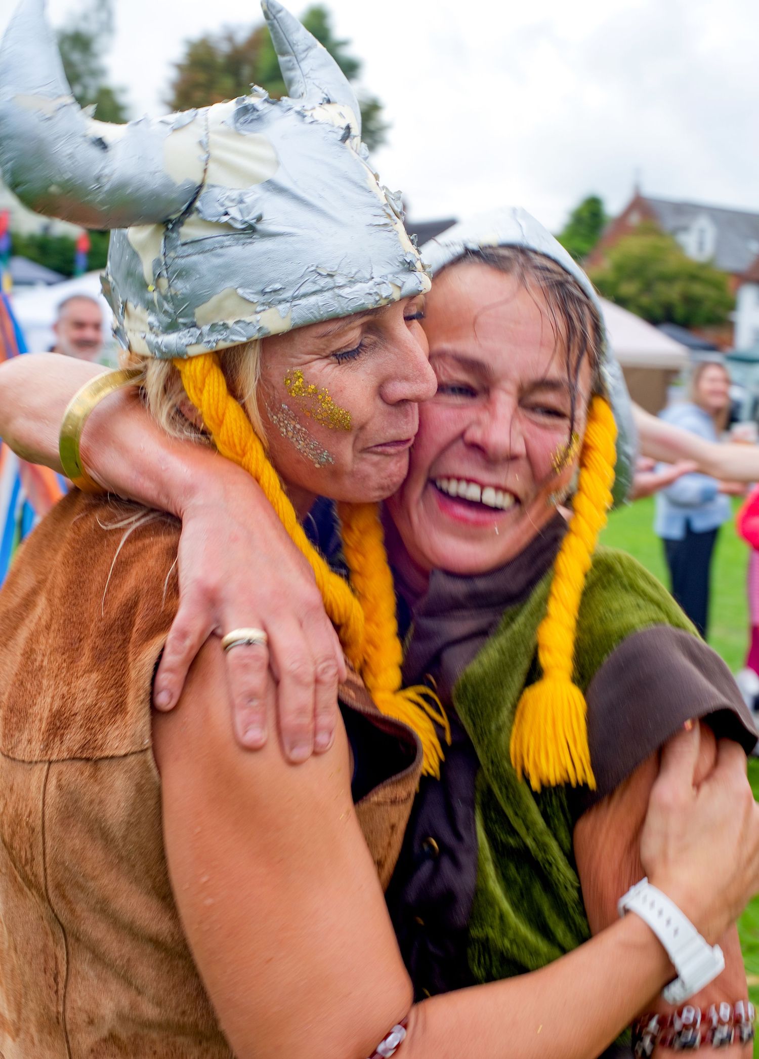 Two women in Viking costumes with yellow braids embrace and laugh at an outdoor festival event.