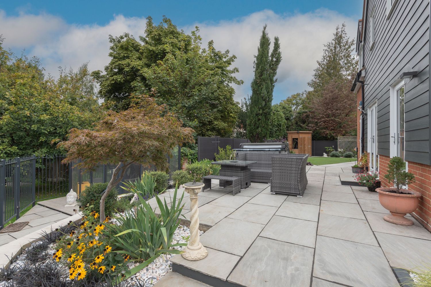 Spacious patio with grey stone tiles, outdoor seating, ornamental Japanese maple, yellow flowers, and lush garden backdrop.