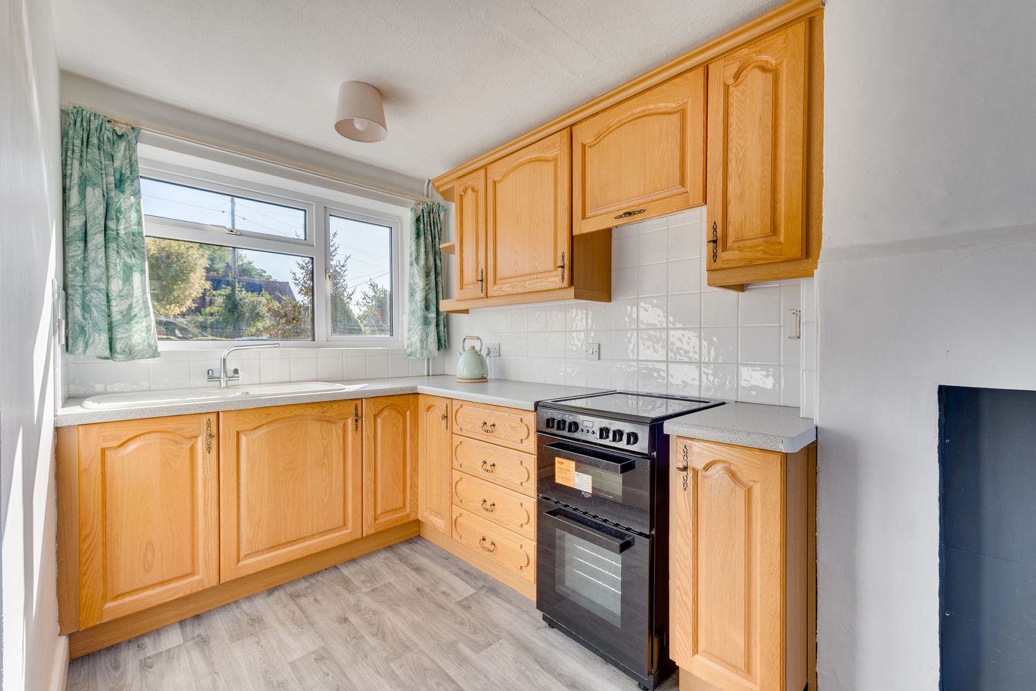 Light wood kitchen with black electric cooker, tiled splashback, and green curtained window over sink.