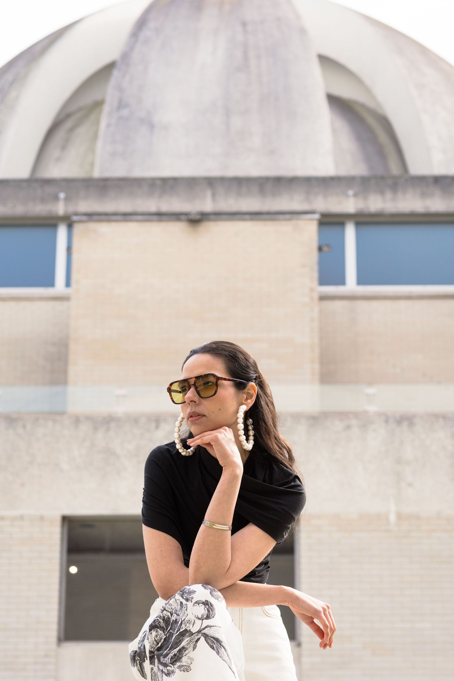 Stylish woman in black top, floral skirt, pearl earrings, and sunglasses posing before a concrete domed building.