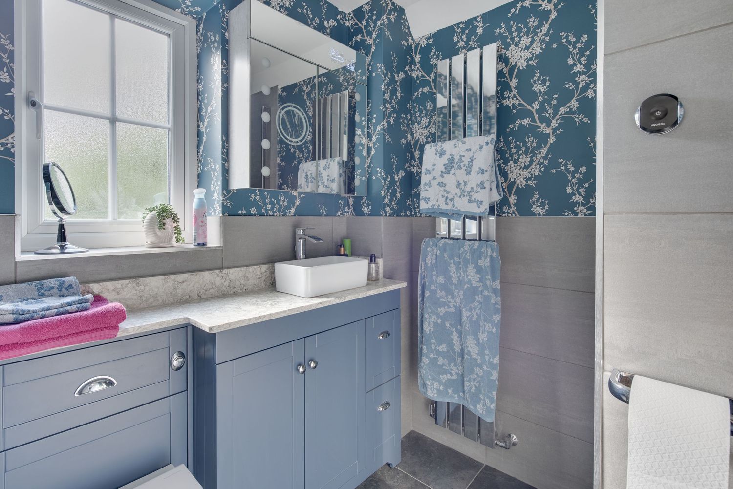 Elegant blue bathroom with floral wallpaper, grey cabinets, vessel sink, large mirror, and tiled shower area.