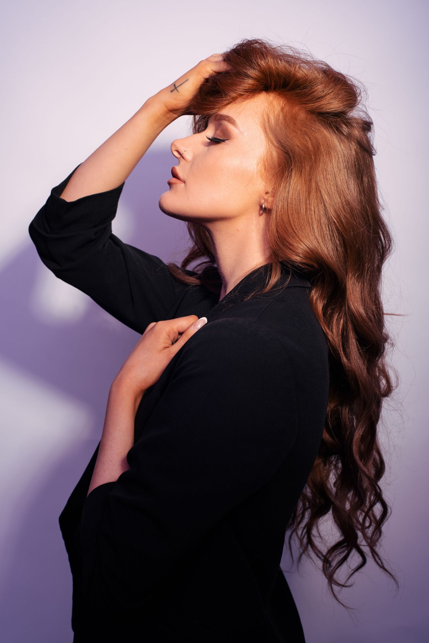 A woman with long auburn wavy hair poses in a black top against a purple background, hand raised to her head.