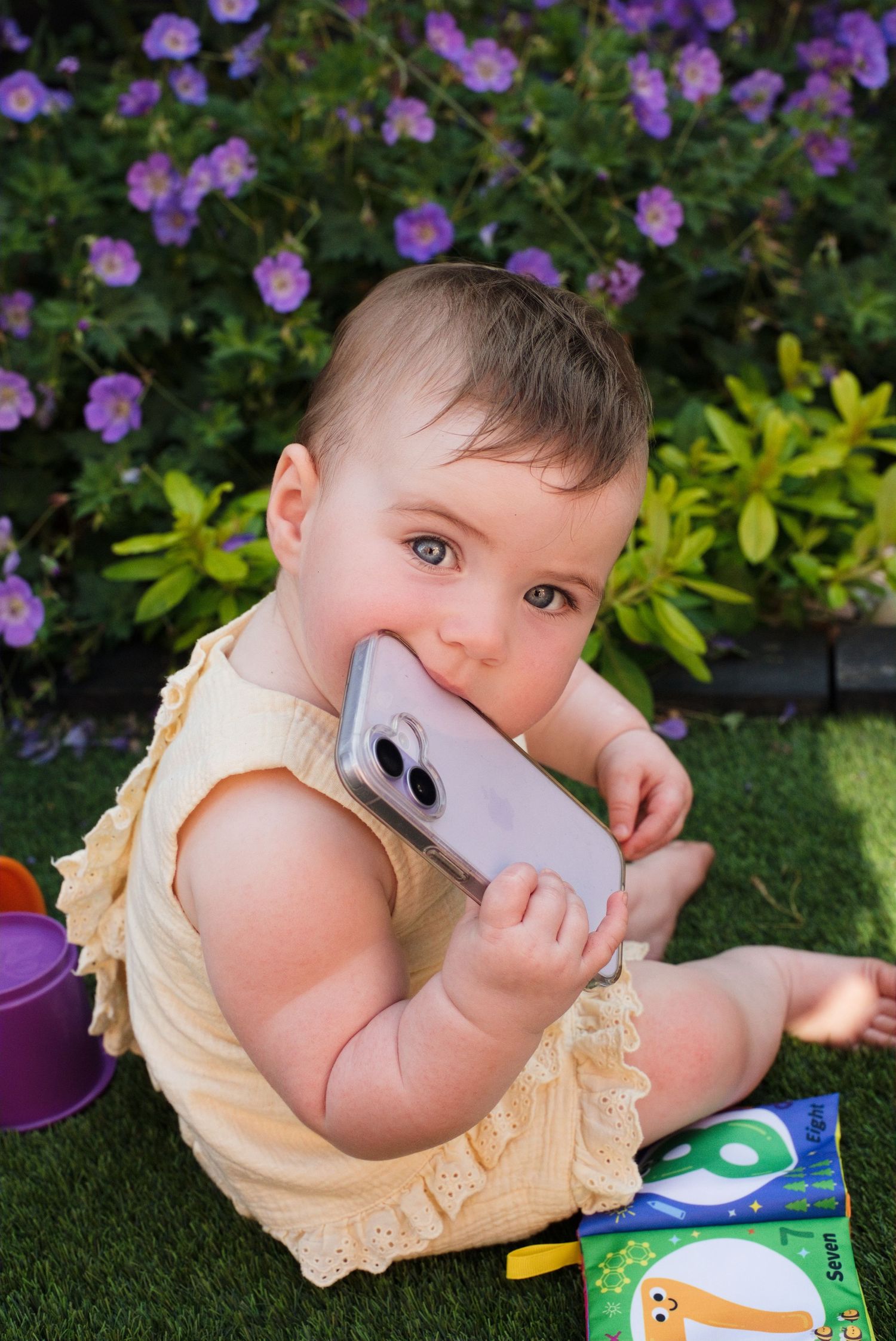 Adorable baby in yellow romper chewing on a smartphone while sitting on grass near purple flowers outdoors.
