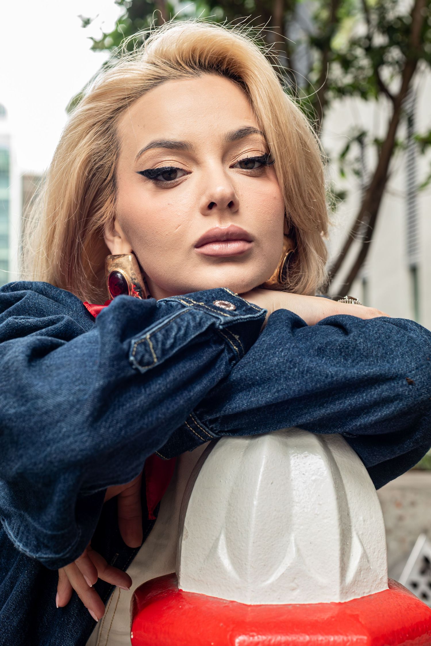 Blonde woman in denim jacket leans on bollard with bold eye makeup outdoors.