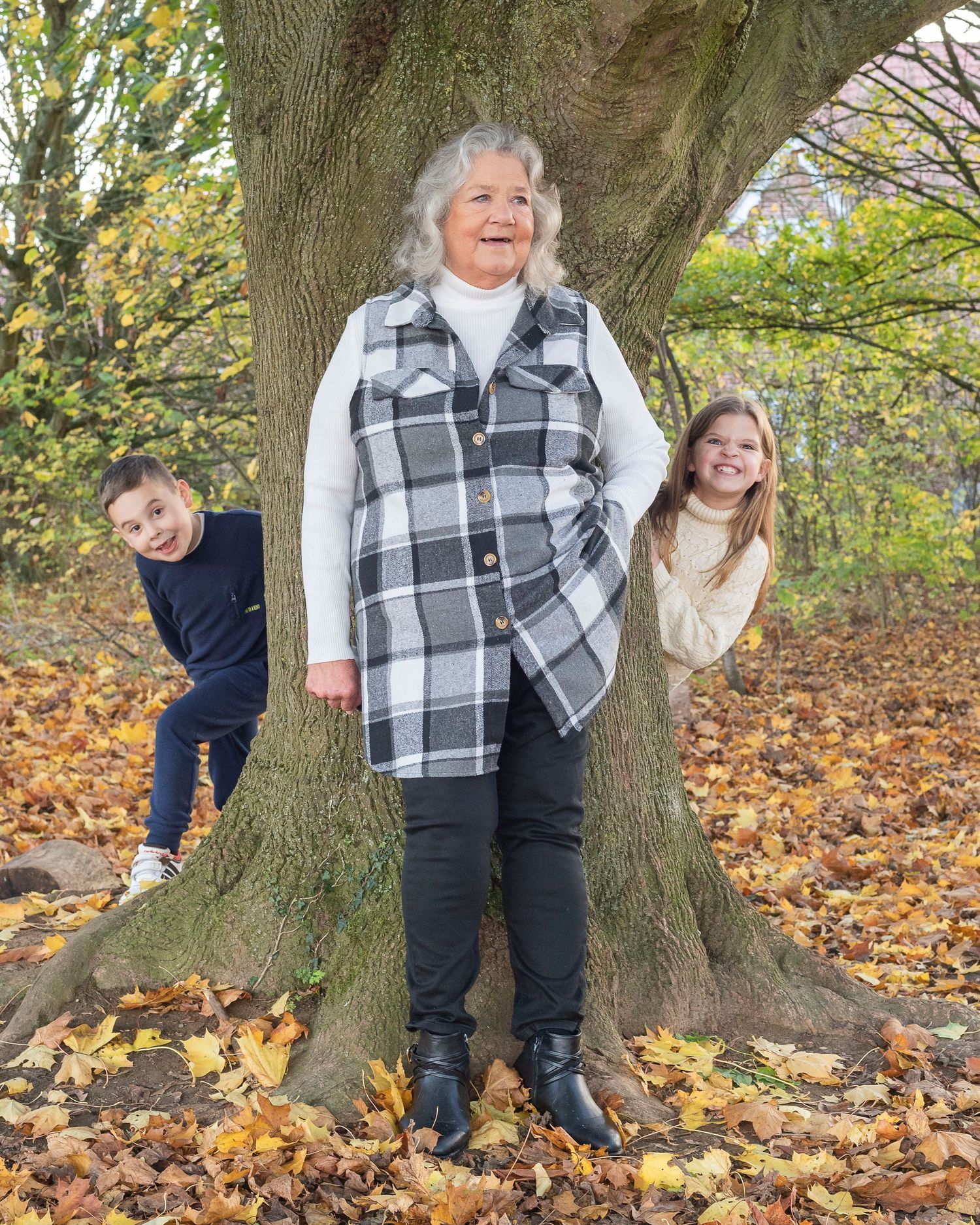 Elderly woman smiling in autumn park with two children peeking from behind a large tree, surrounded by fallen leaves.