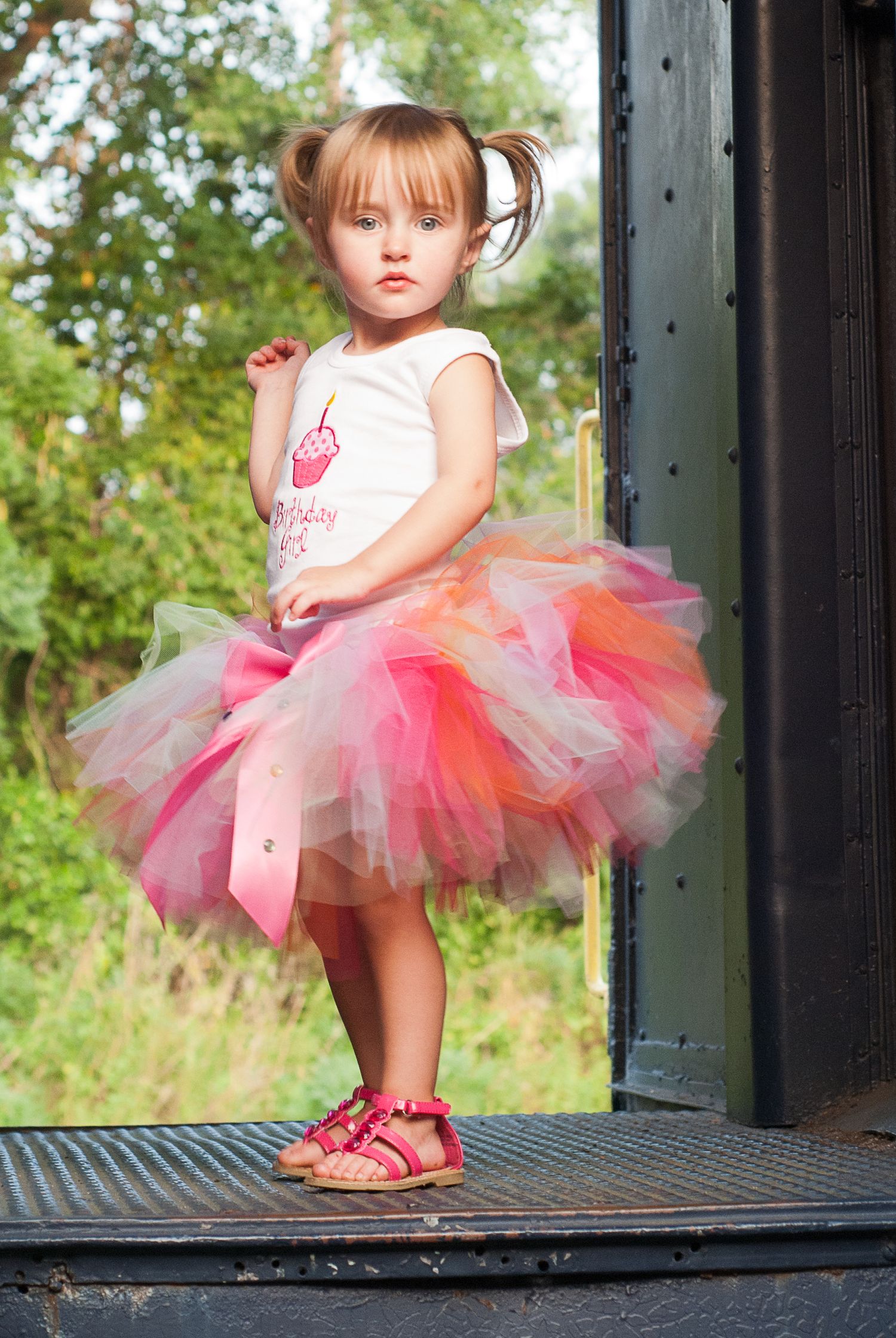 Pretty little girl in birthday tutu outfit standing on metal steps posing with serious expression.