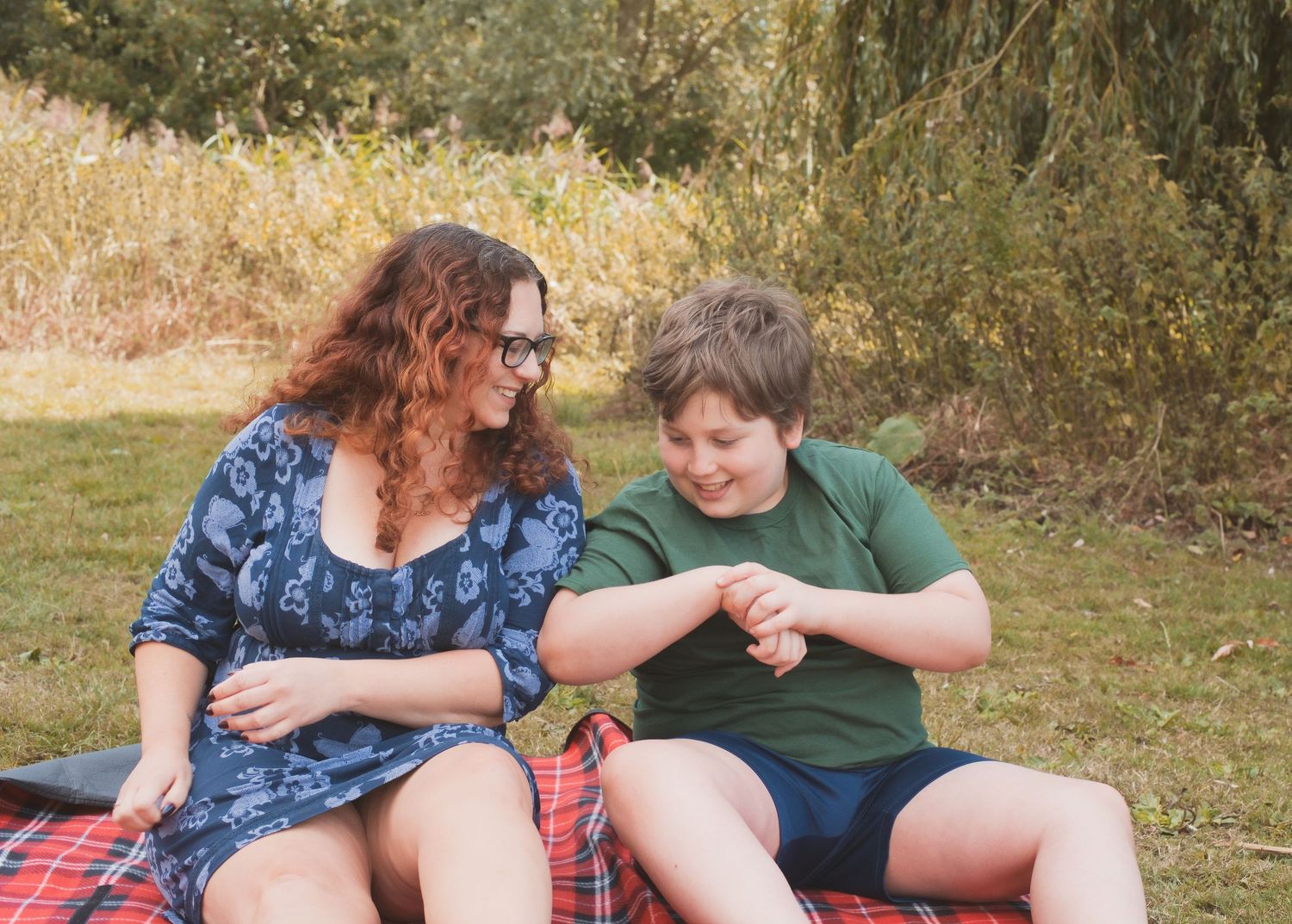 A woman and boy sit on a red blanket outdoors, laughing together in a sunny park setting.