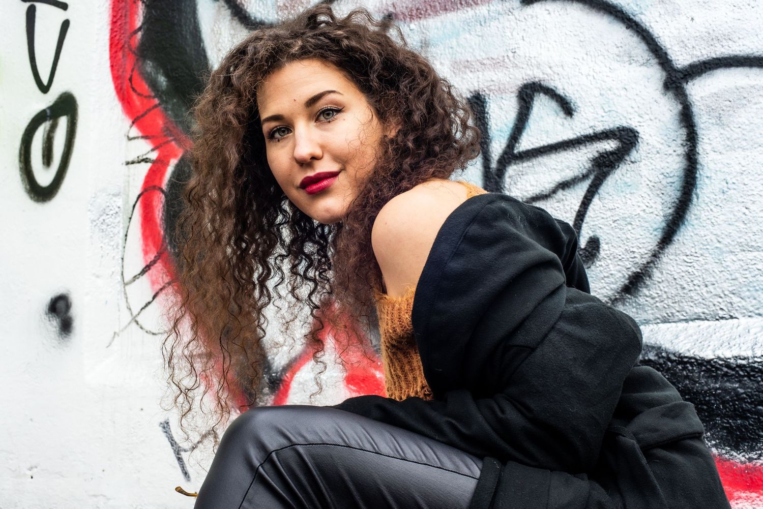 Young woman with curly hair in black outfit posing against a colorful graffiti wall outdoors.