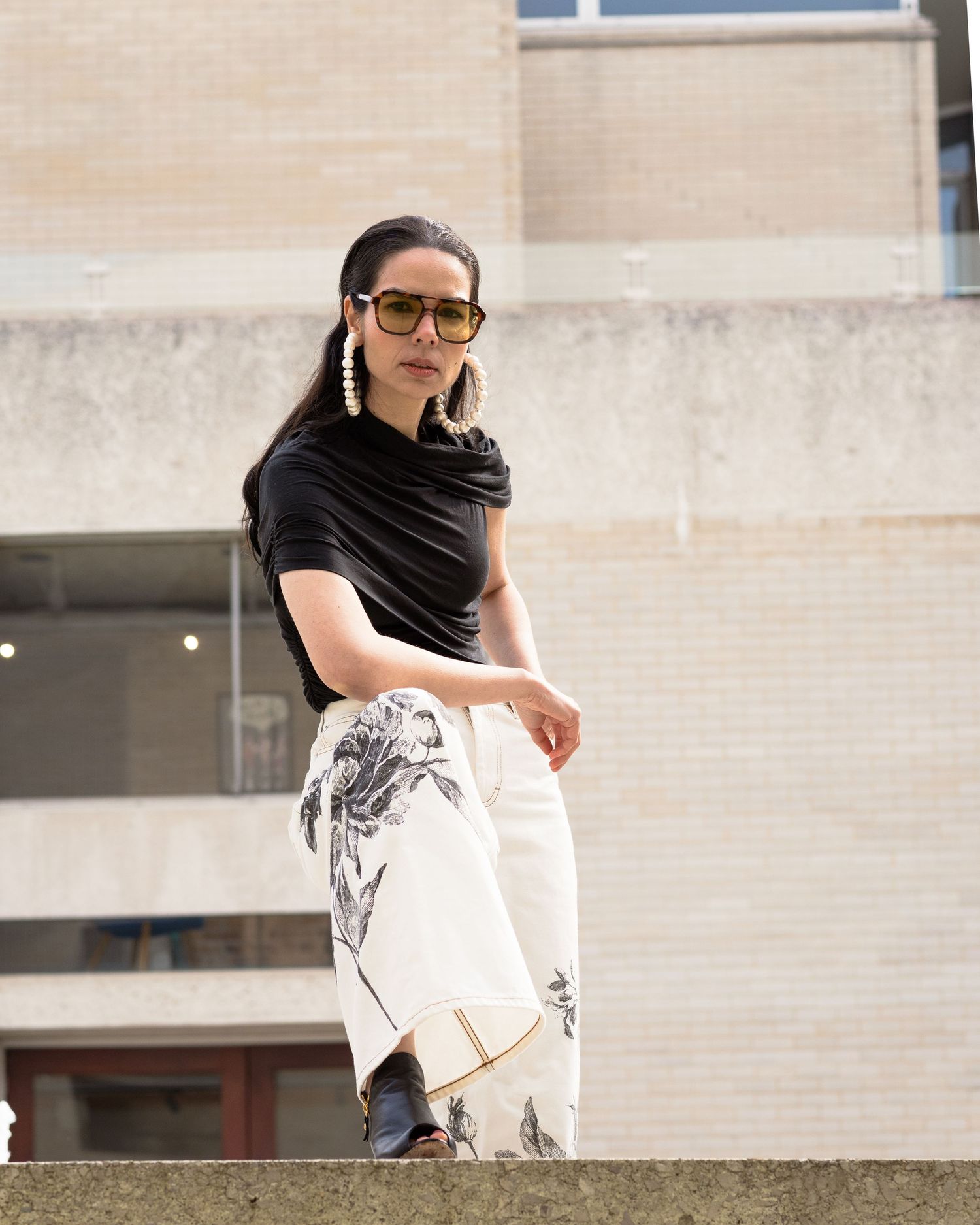 Woman in black draped top and white floral printed trousers poses outdoors wearing sunglasses and pearl earrings.