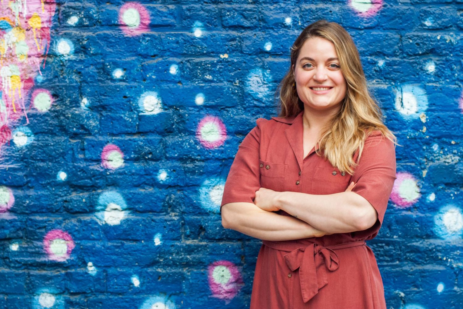 Smiling woman in rust dress poses arms crossed against vibrant blue graffiti mural wall.