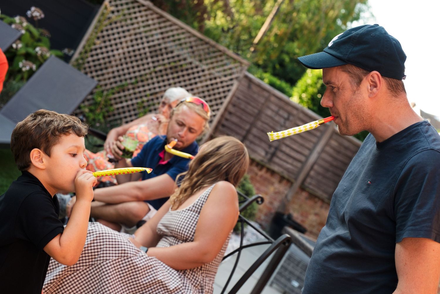 Family and friends enjoy a fun outdoor gathering, playfully blowing on yellow party horns in a garden setting.