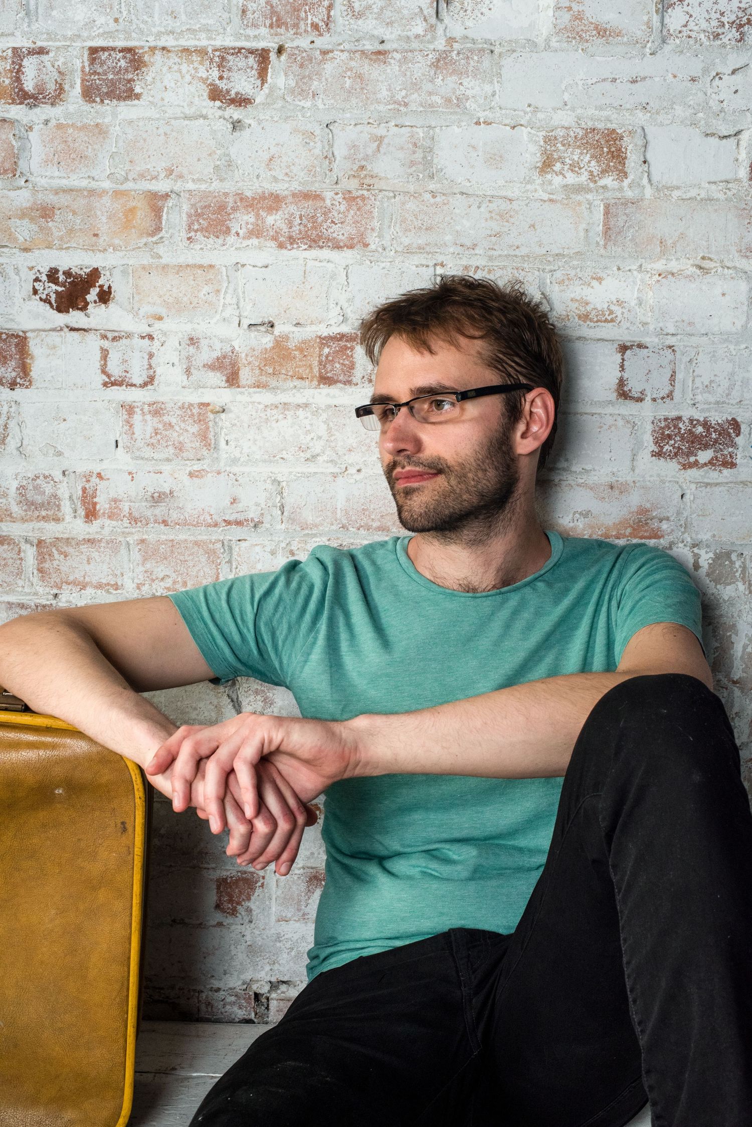 Man in teal t-shirt and glasses sitting relaxed against a white brick wall.