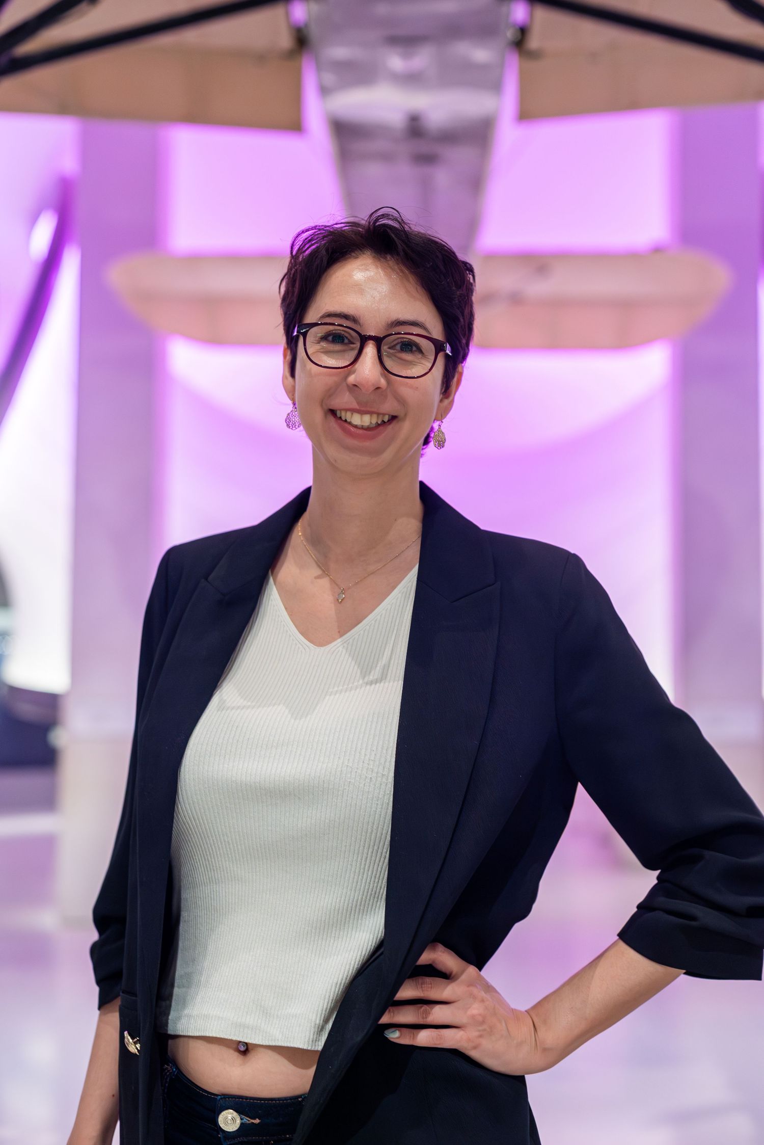 Woman in black blazer and white top smiling confidently in a modern purple-lit interior space.