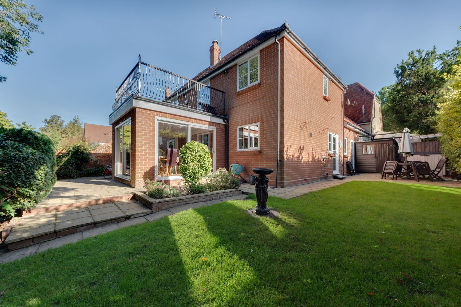 Rear garden view of a brick detached house with balcony, glass extension, patio area, and well-maintained lawn surrounded by mature trees.