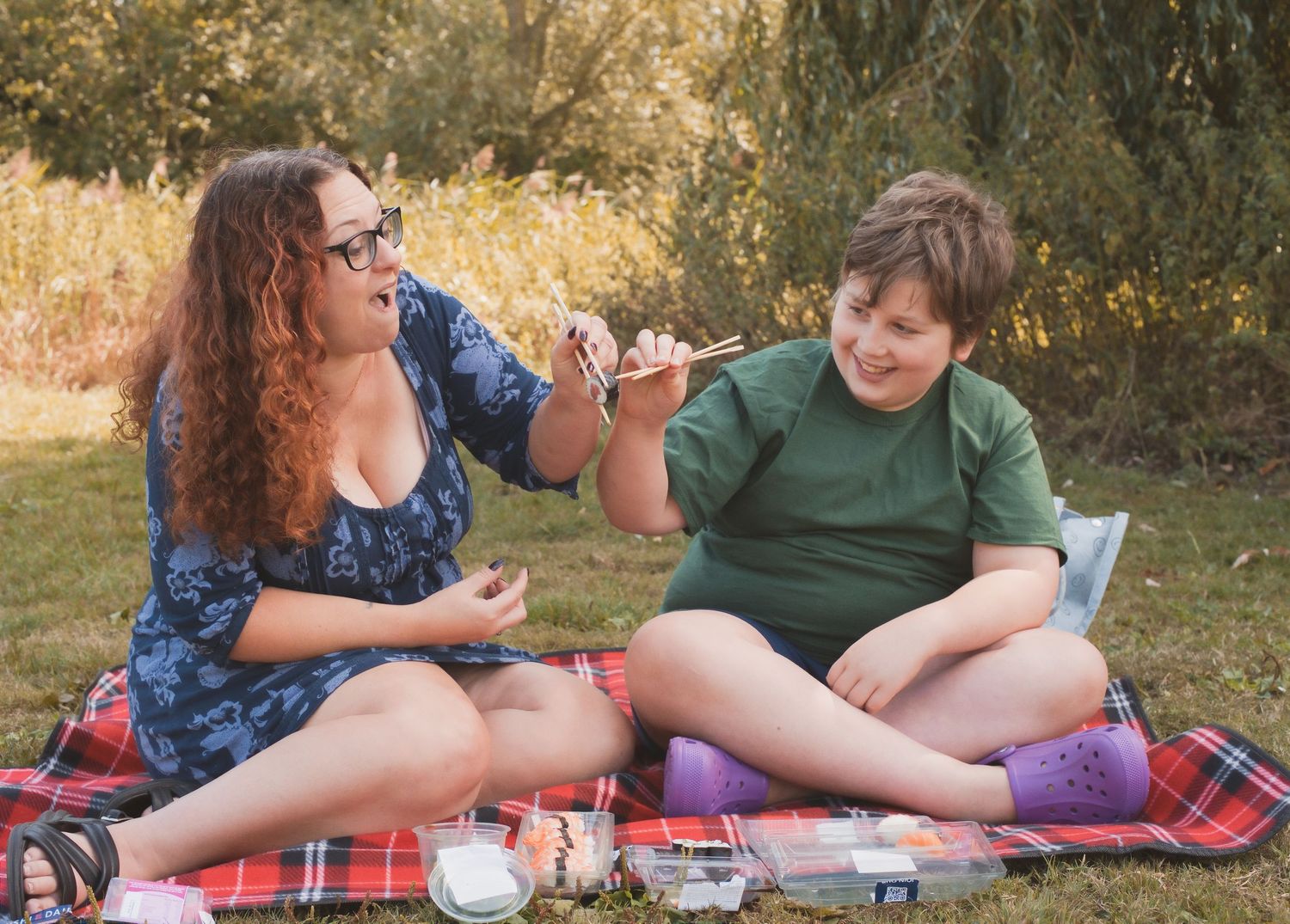 Two friends enjoy a picnic on a plaid blanket outdoors, sharing food and smiling on a sunny day.