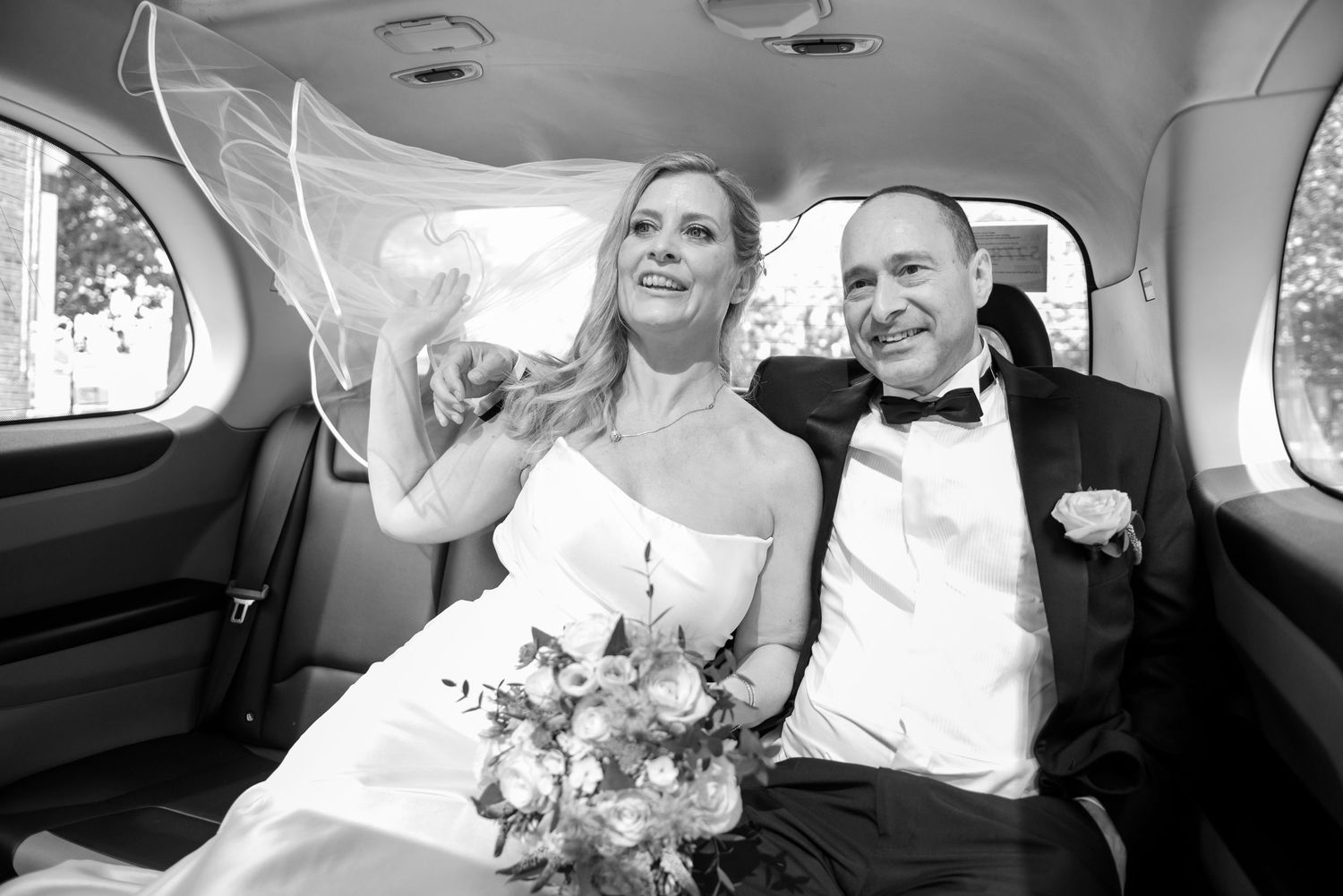 Black and white photo of a smiling bride and groom sitting together in a car on their wedding day.