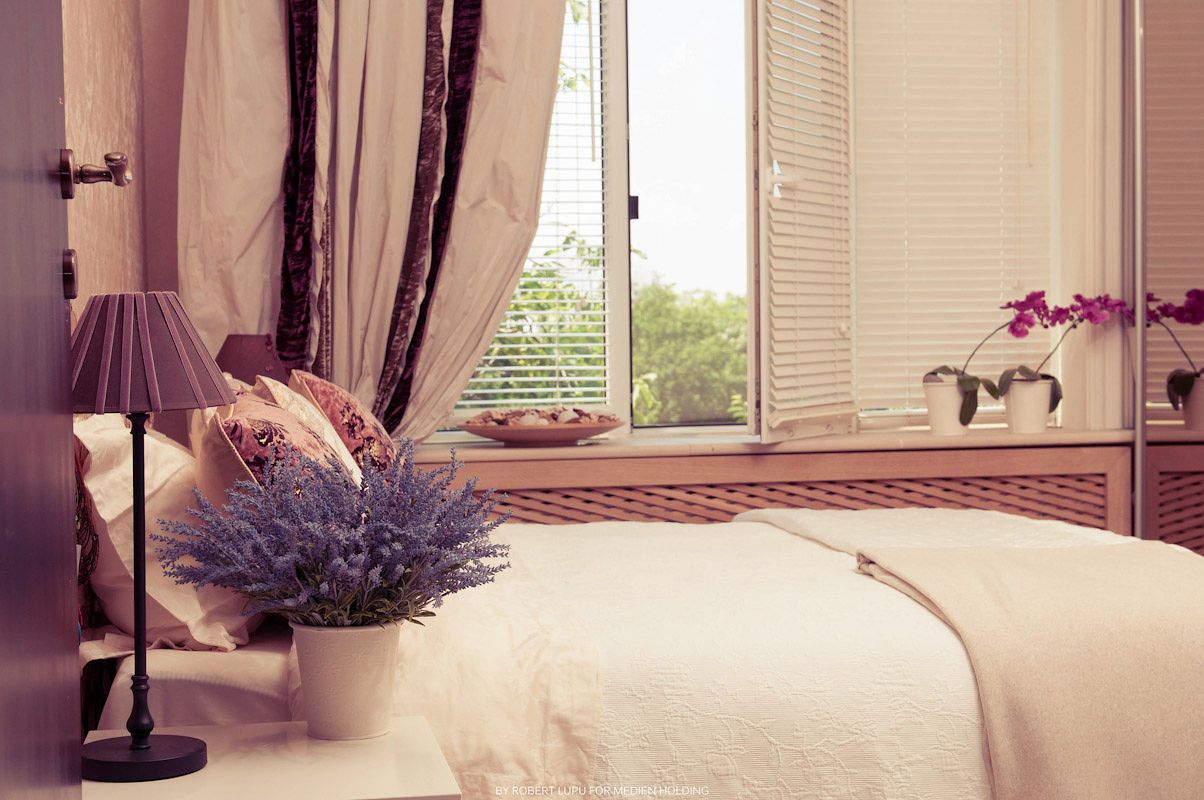 Cozy bedroom with white bedding, lavender flowers, purple lamp, sheer curtains, and bright window with orchids on sill.