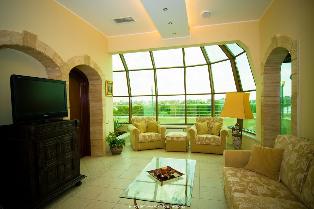 Elegant hotel suite living room with arched windows, glass coffee table, floral chairs, and flat-screen TV.