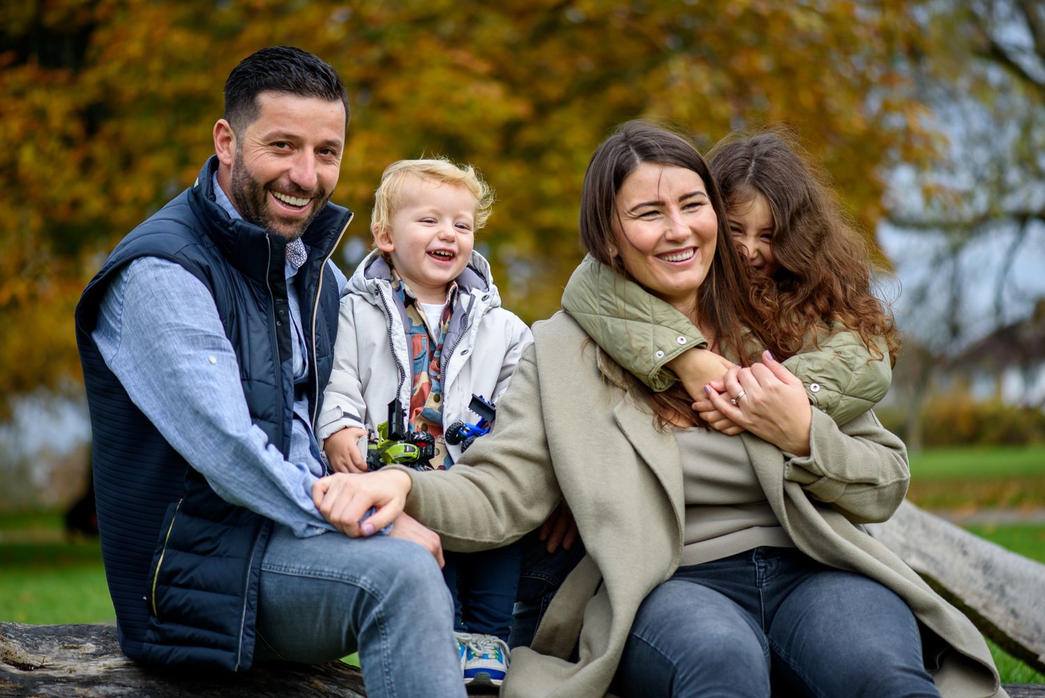 Happy family of four sitting outdoors on a log in an autumn park, smiling and embracing on a cool fall day.