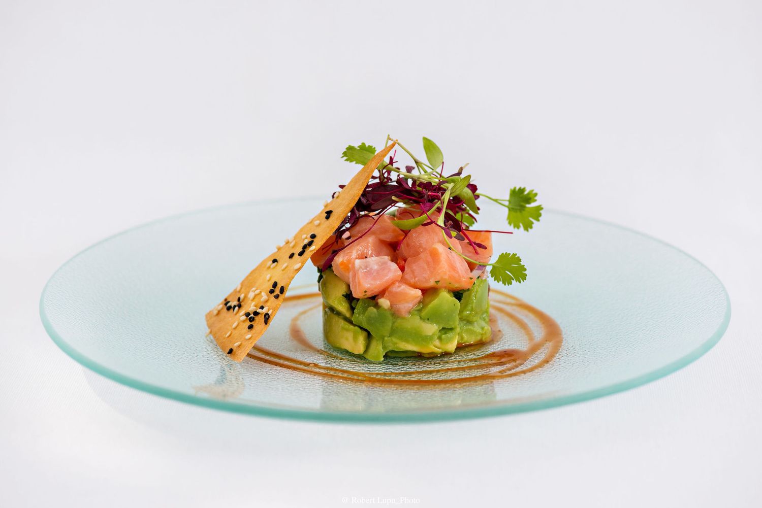 Elegant salmon tartare with avocado, microgreens, and sesame crisp on a glass plate, fine dining presentation.