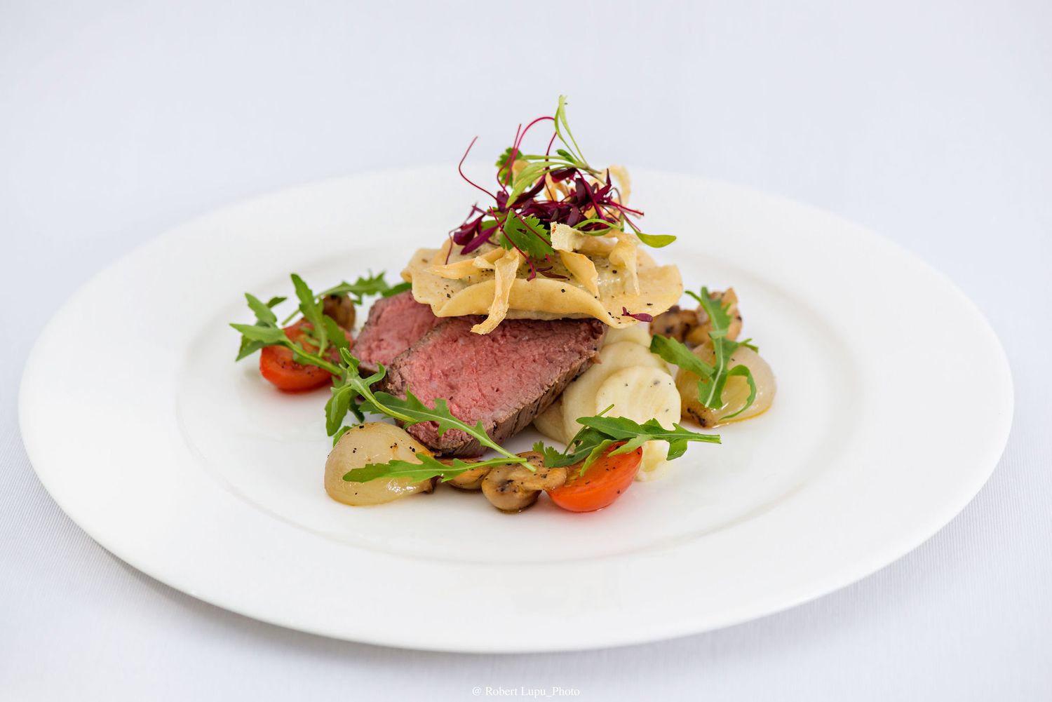 Elegant plated dish featuring sliced medium-rare beef on sautéed vegetables, topped with crispy onions and fresh microgreens.