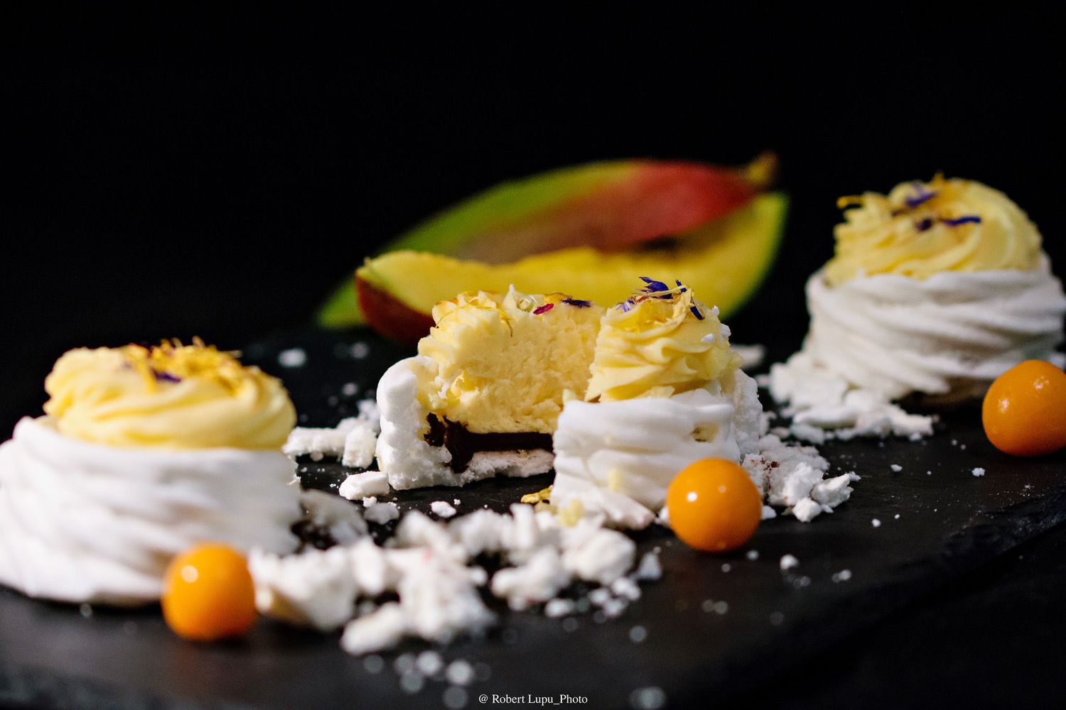 Elegant meringue desserts topped with lemon cream and coconut flakes, served with mango slices and golden berries on a dark slate.