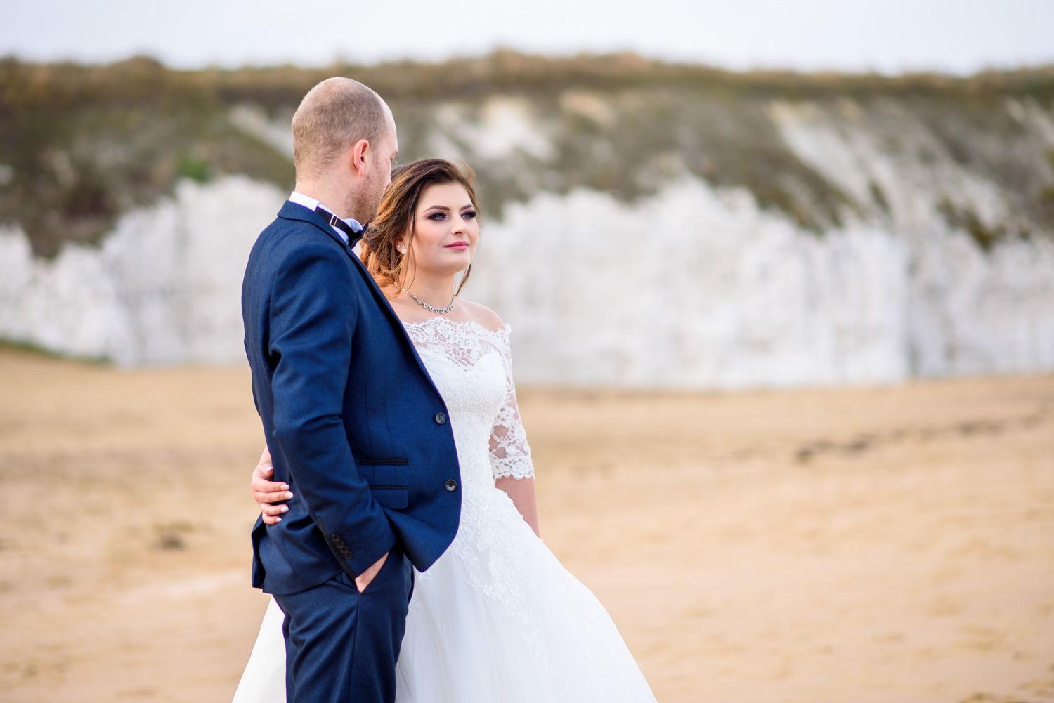 Bride and groom embracing on a sandy beach with white chalk cliffs in the background on their wedding day.