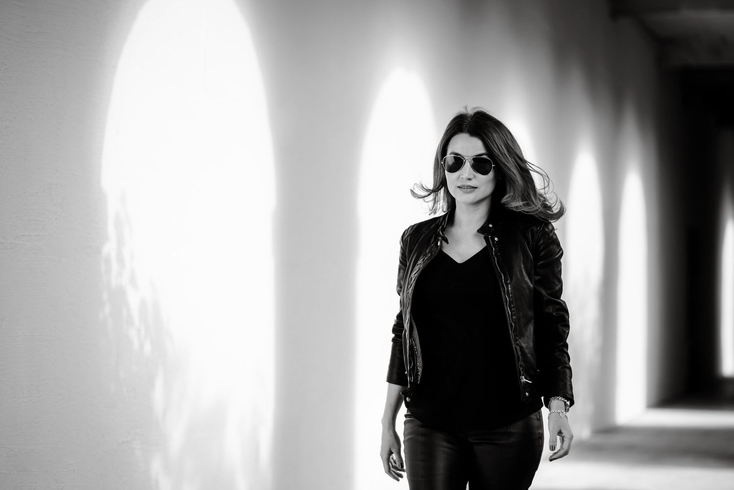 Woman in leather jacket and sunglasses walking through arched corridor in black and white photo.