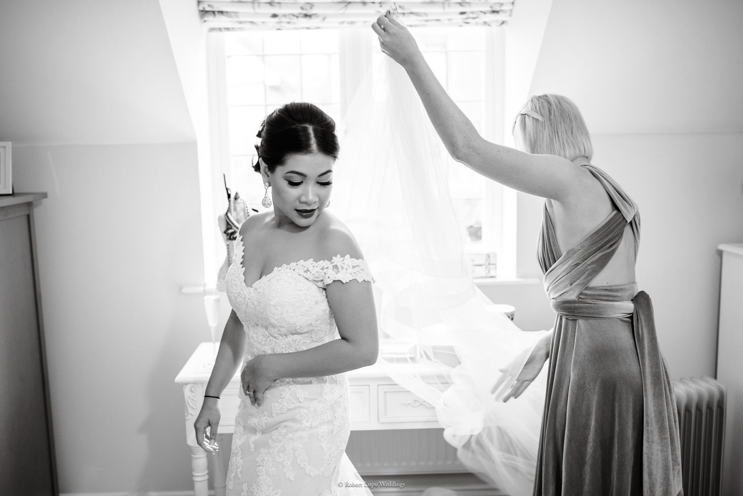 Black and white photo of a bride in lace gown being helped by bridesmaid near bright window.