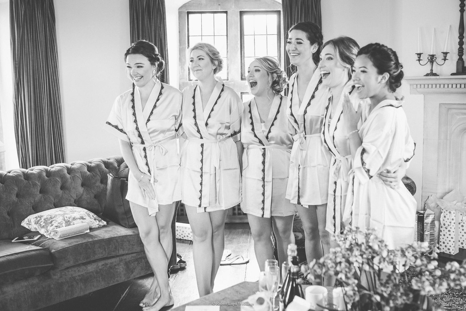 Bridesmaids in matching robes laughing together in elegant room during wedding preparation, black and white photo.