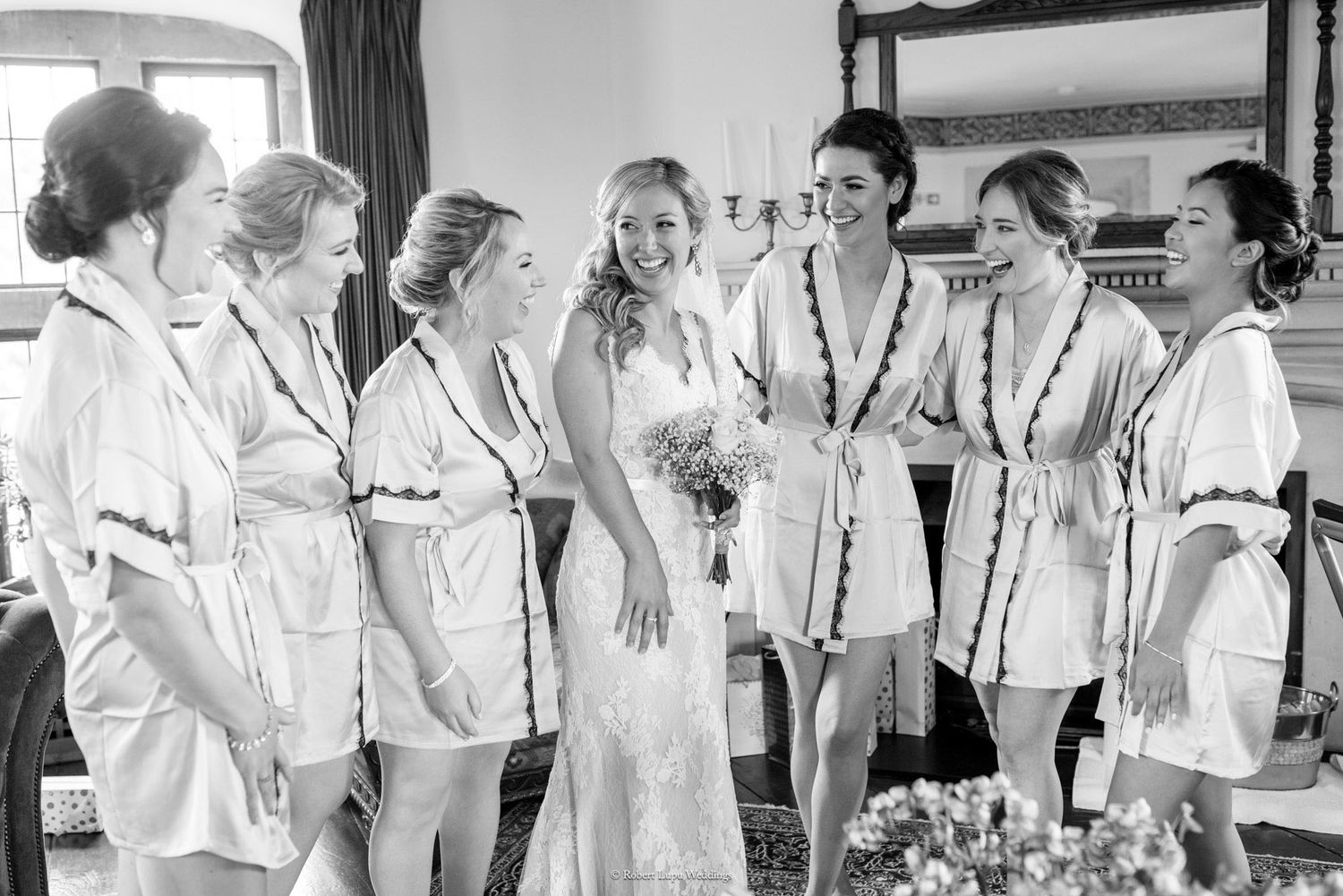 Bride in lace gown smiles with bridesmaids in matching robes during wedding morning preparations.