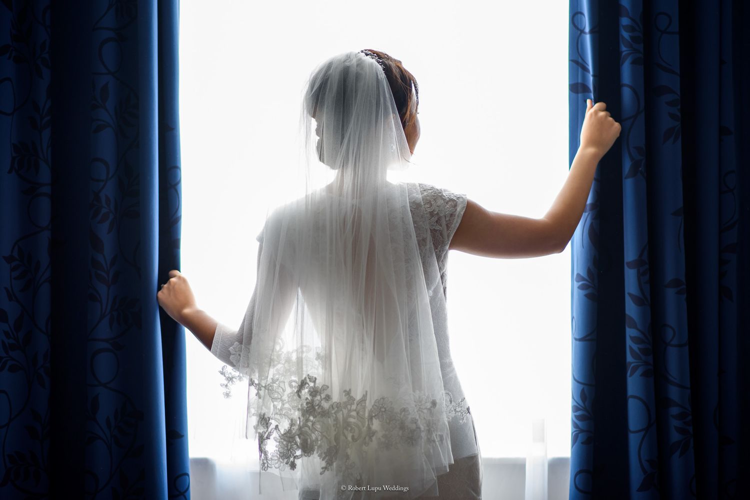 Bride in wedding dress and veil stands at blue curtained window holding drapes open, backlit by natural light.