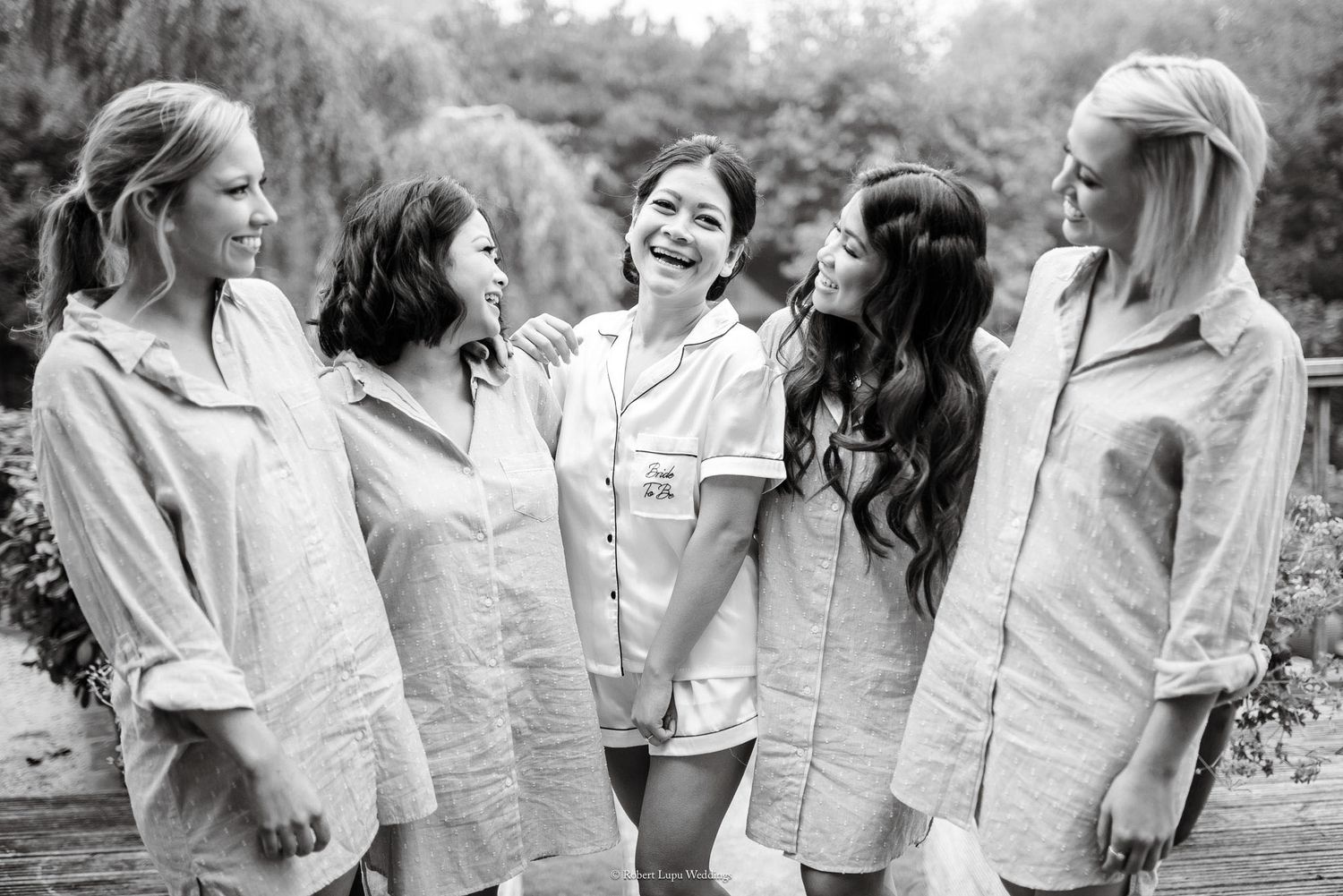 Five women in matching linen shirts laughing together outdoors in a black and white bridal party photo.