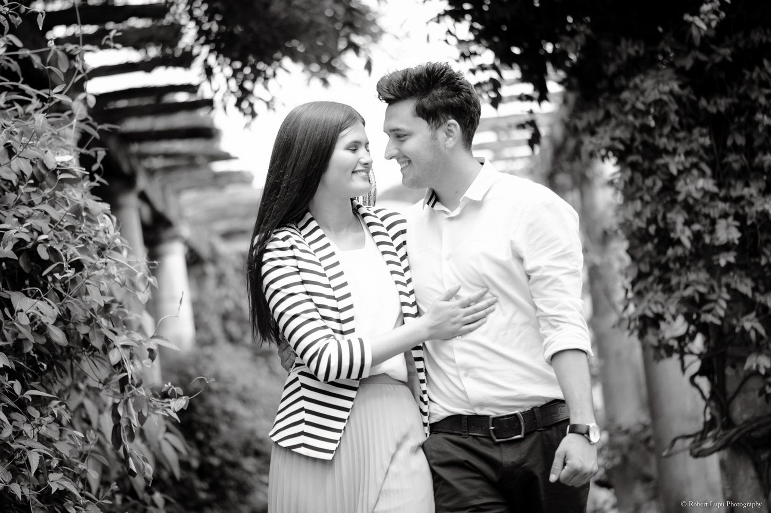 Black and white photo of a smiling couple sharing a romantic moment outdoors among lush ivy-covered walls.