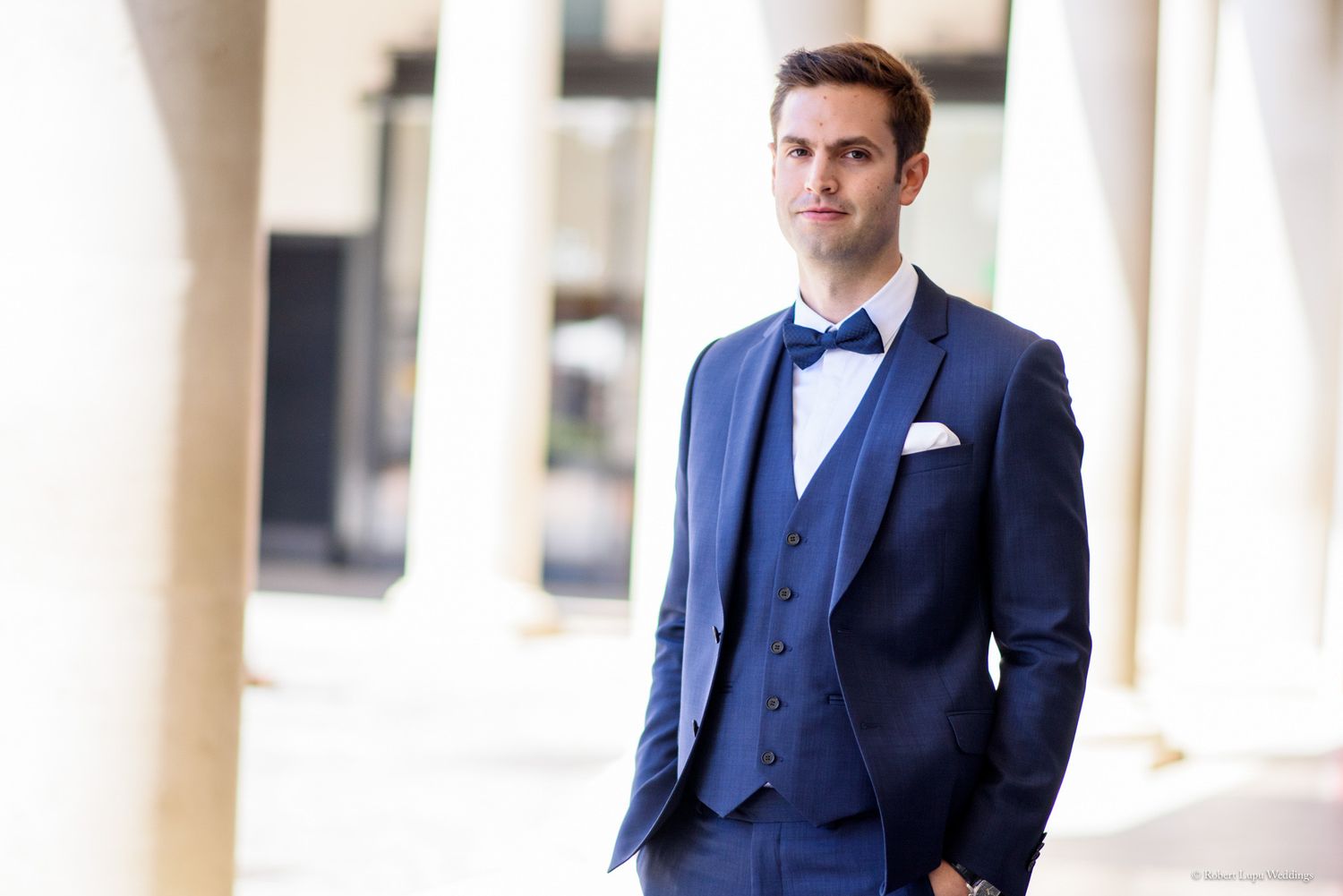 Elegant man in a navy blue three-piece suit with bow tie and pocket square posing outdoors.