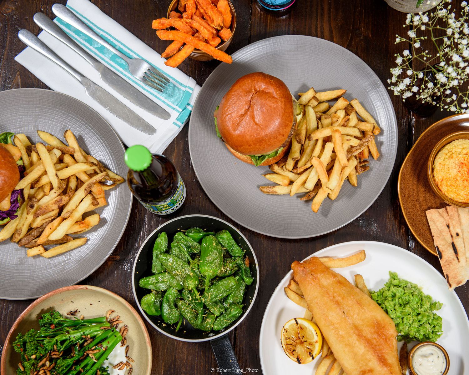 Overhead view of restaurant table with burger, fries, fish and chips, greens, and sweet potato fries on plates.