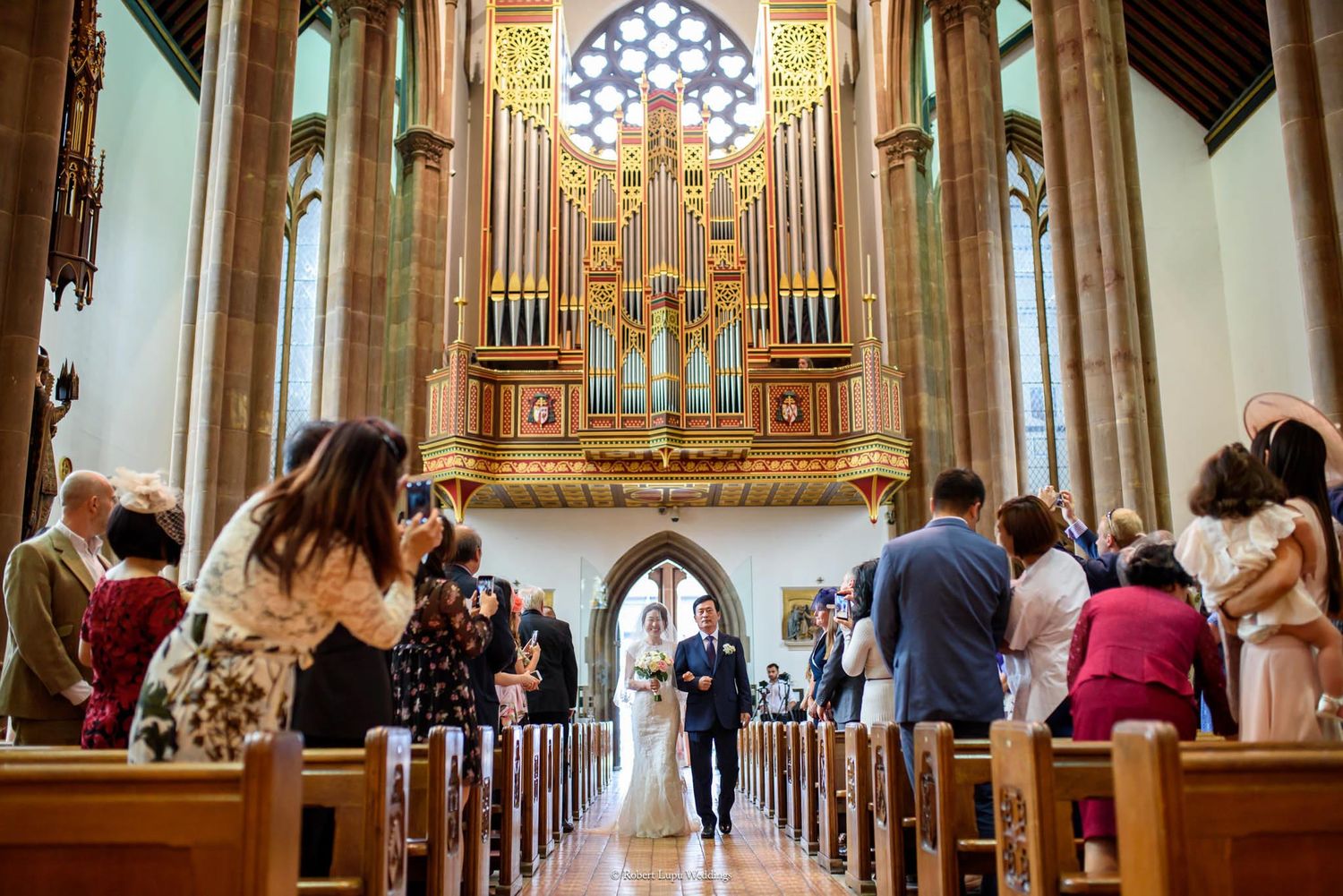 Bride and groom walk down the aisle of a grand church with a large pipe organ during their wedding ceremony.