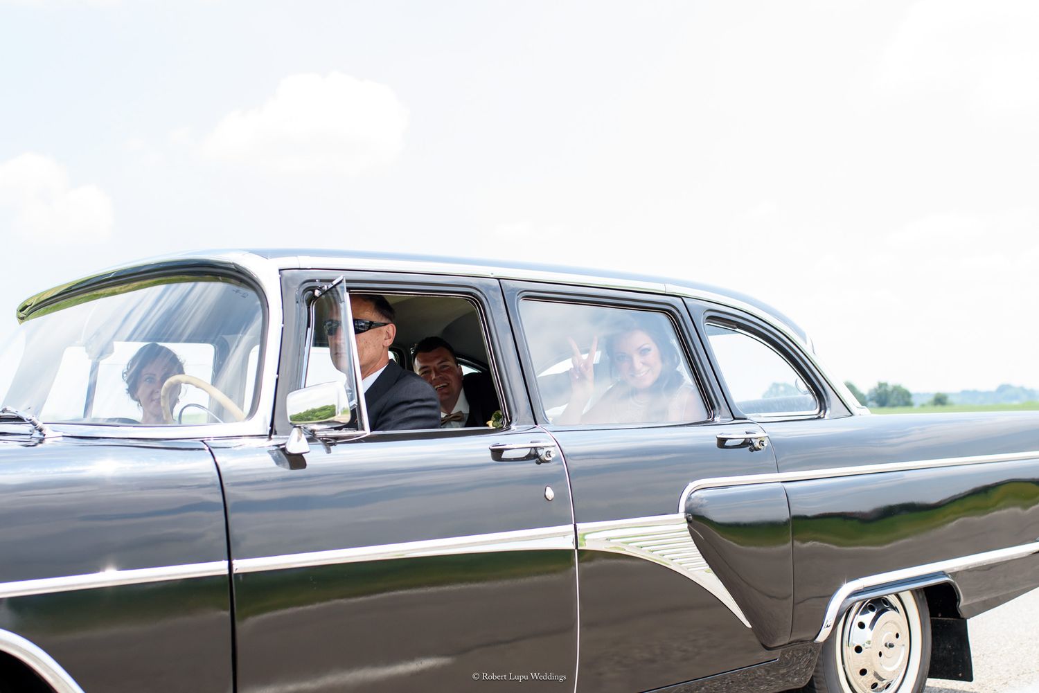 Vintage black classic car with passengers visible through windows, parked on a bright sunny day.