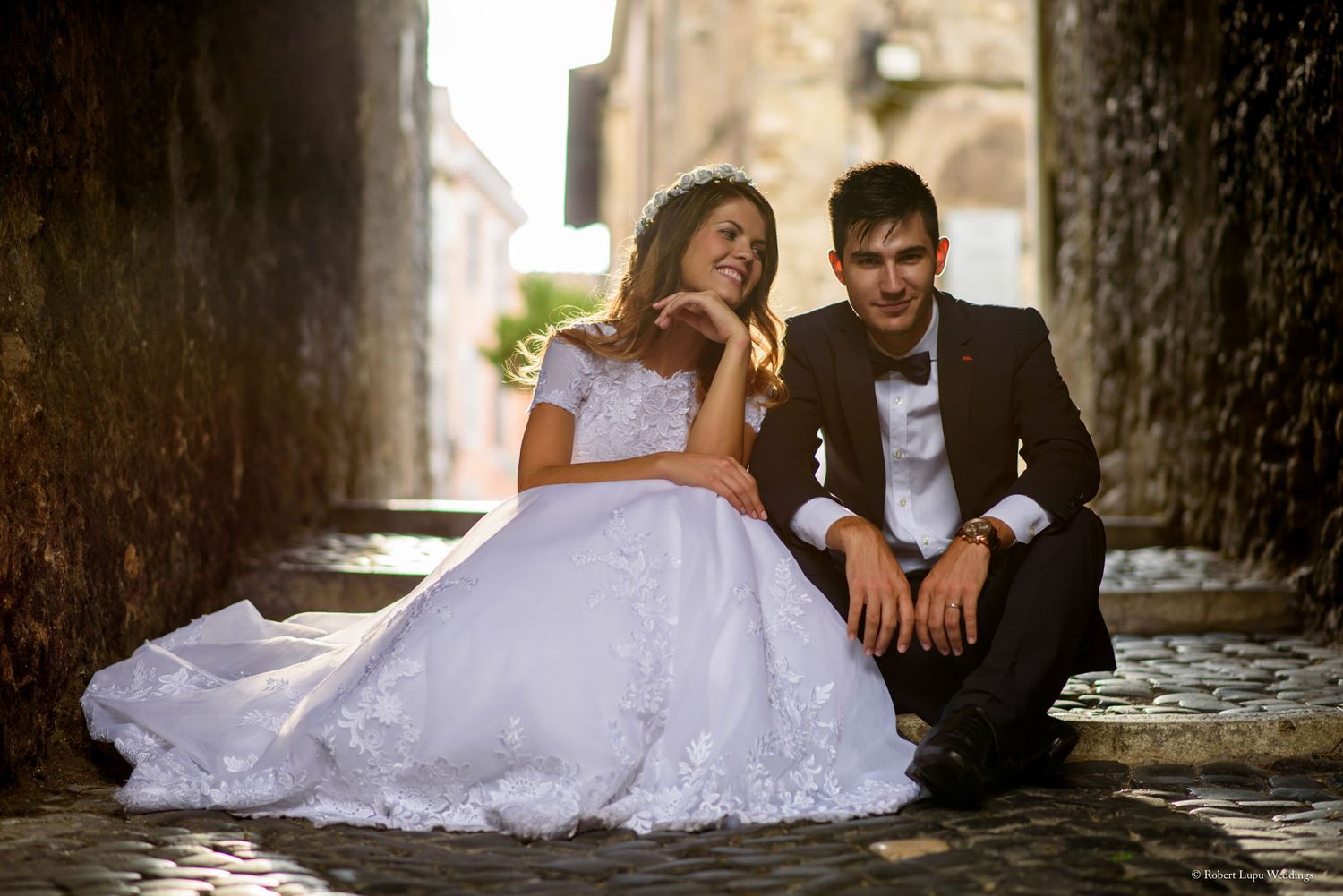 Bride in white lace gown and groom sitting together on cobblestone steps smiling.