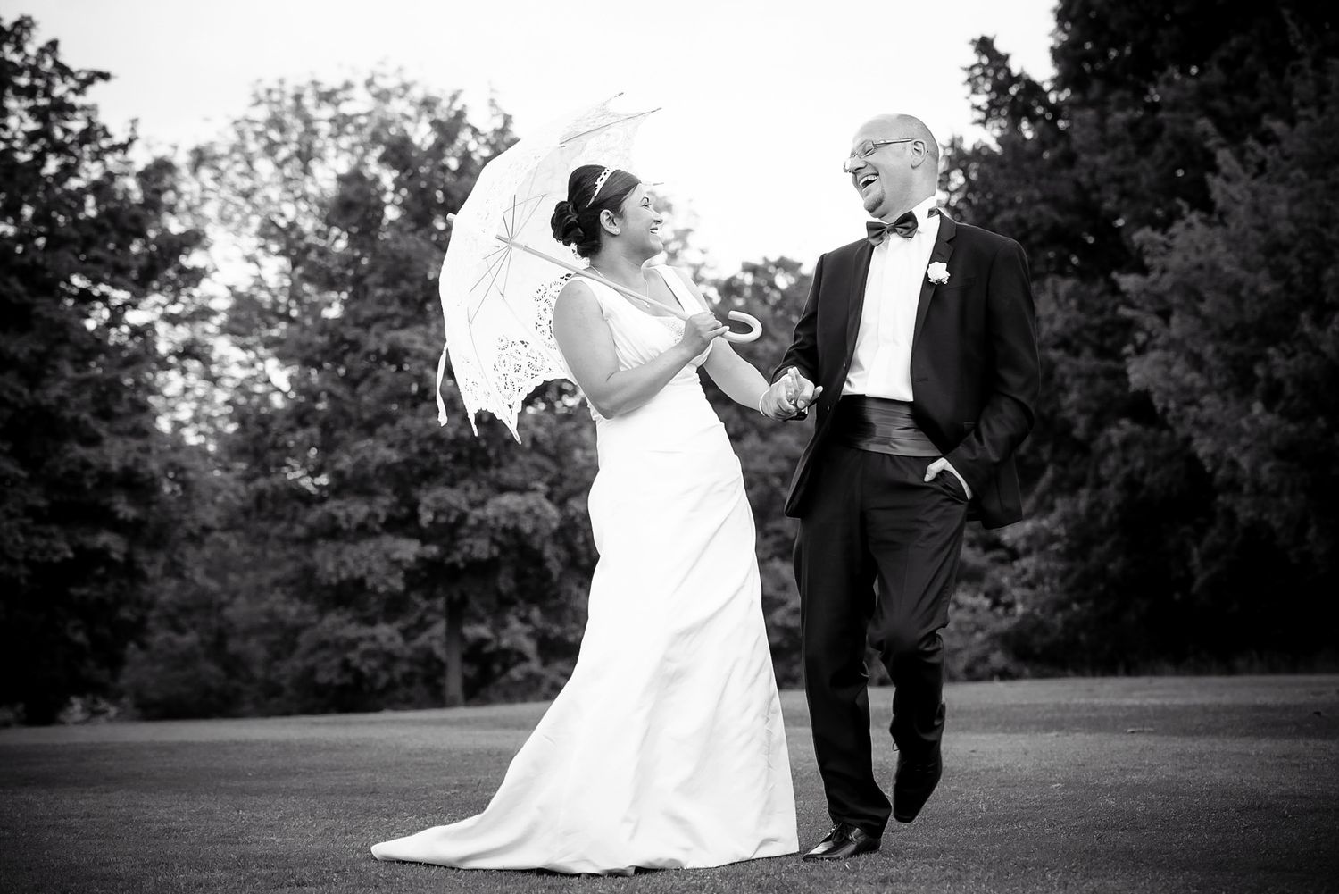 Black and white photo of bride and groom walking outdoors, bride holding a lace parasol umbrella.