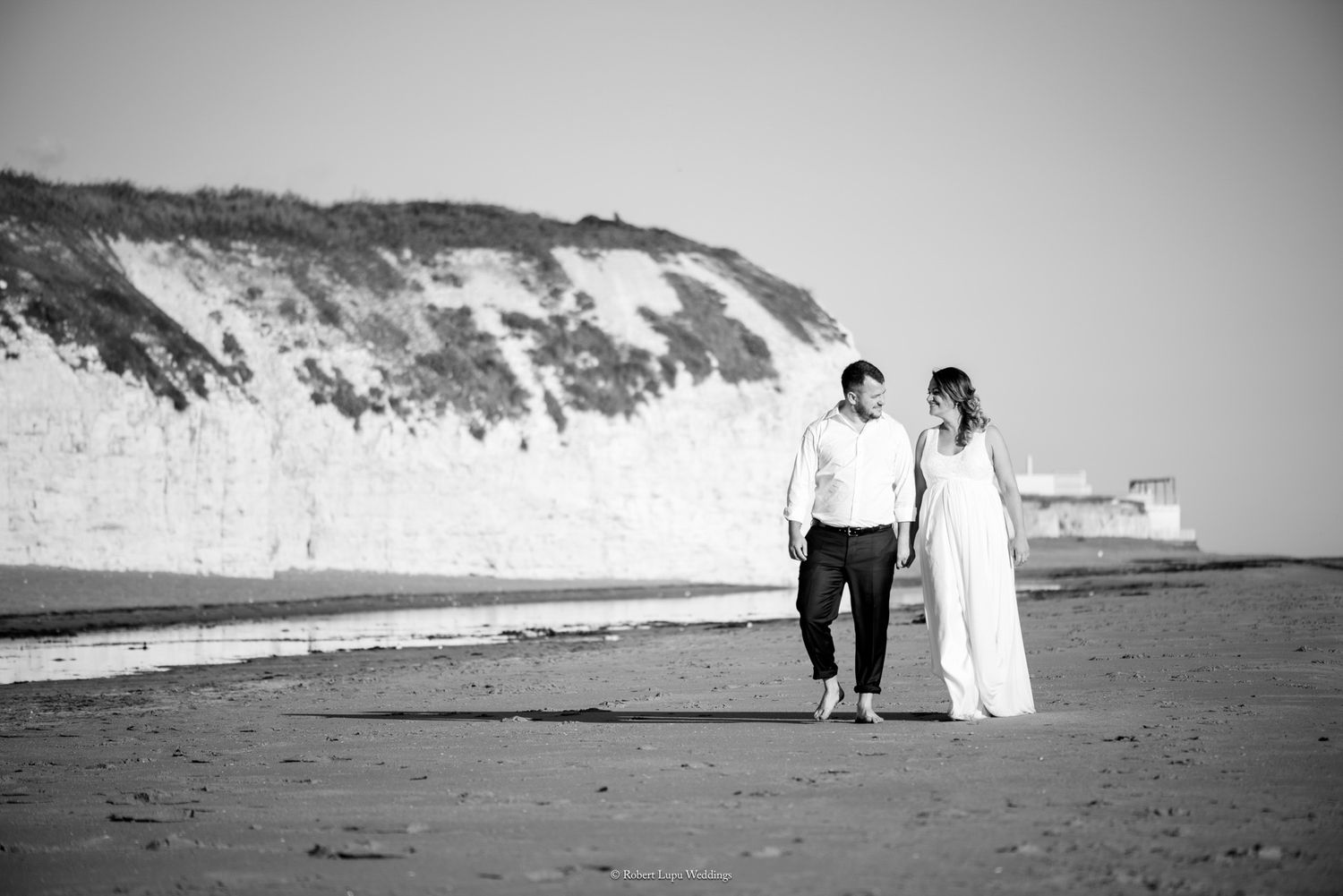 Black and white photo of a couple in wedding attire walking hand in hand on a beach with white cliffs behind them.