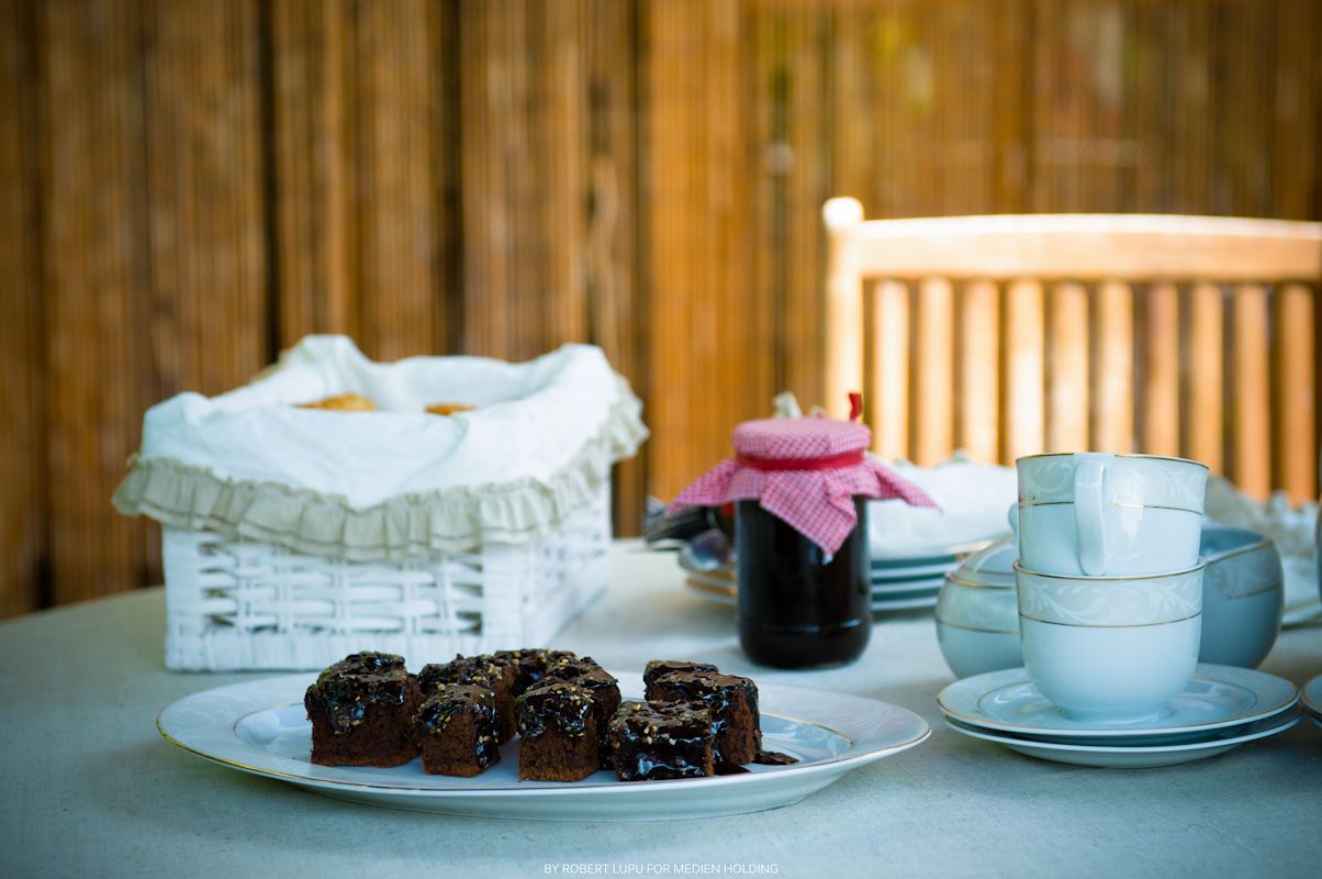 Chocolate brownies on a white plate beside stacked cups, a jar of jam, and a wicker basket on a table.