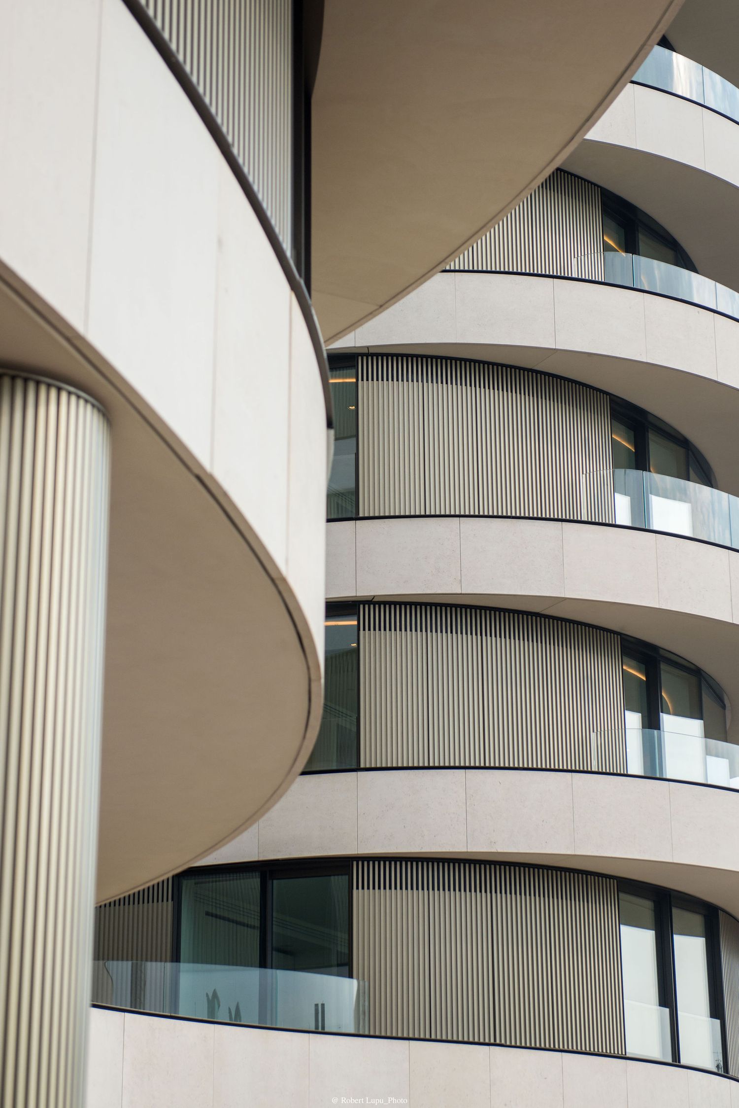 Modern curved building facade with layered balconies, vertical louvers, and floor-to-ceiling windows in neutral tones.