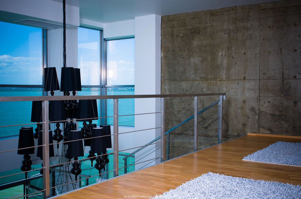 Modern interior balcony with glass railings, hardwood floors, concrete wall, white rug, and ocean view chandelier reflection.