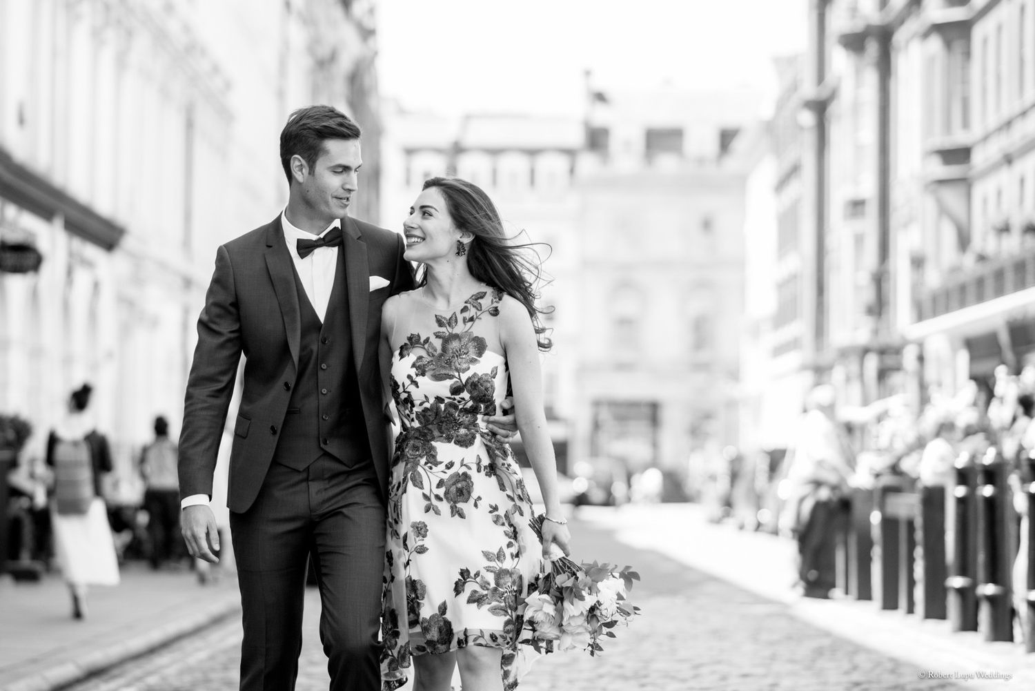 Elegant couple walking on cobblestone street, man in tuxedo and woman in floral dress, black and white wedding photo.