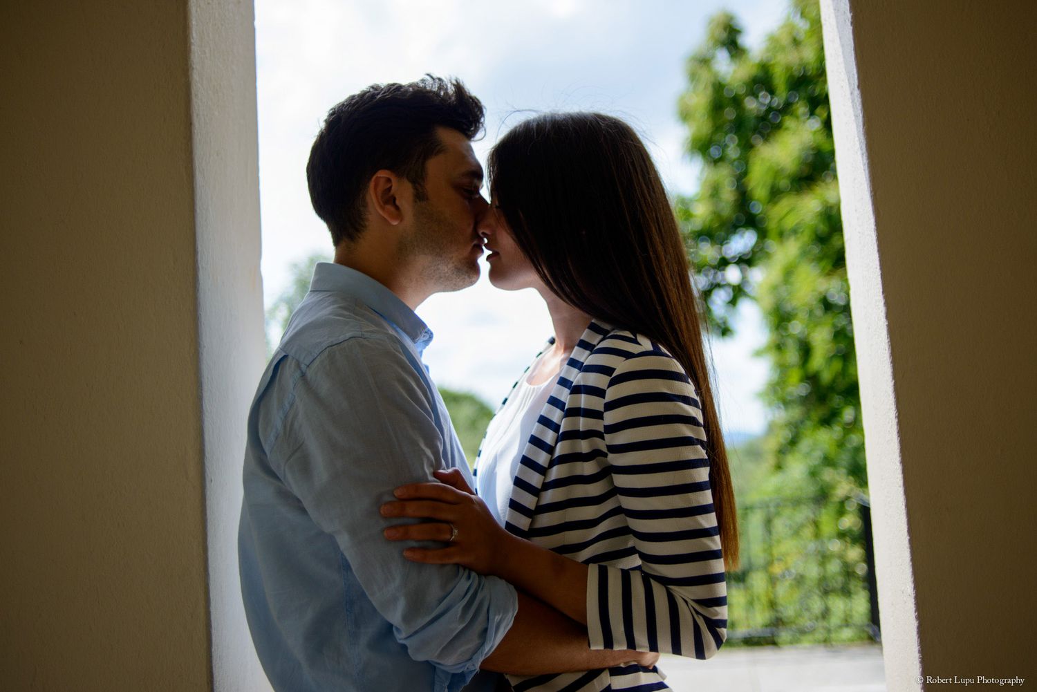 A young couple sharing a romantic kiss near a window with lush green trees in the background.