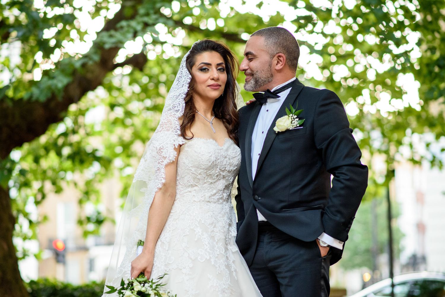 Bride in ornate lace wedding gown with veil and groom in black tuxedo pose together under green leafy trees.