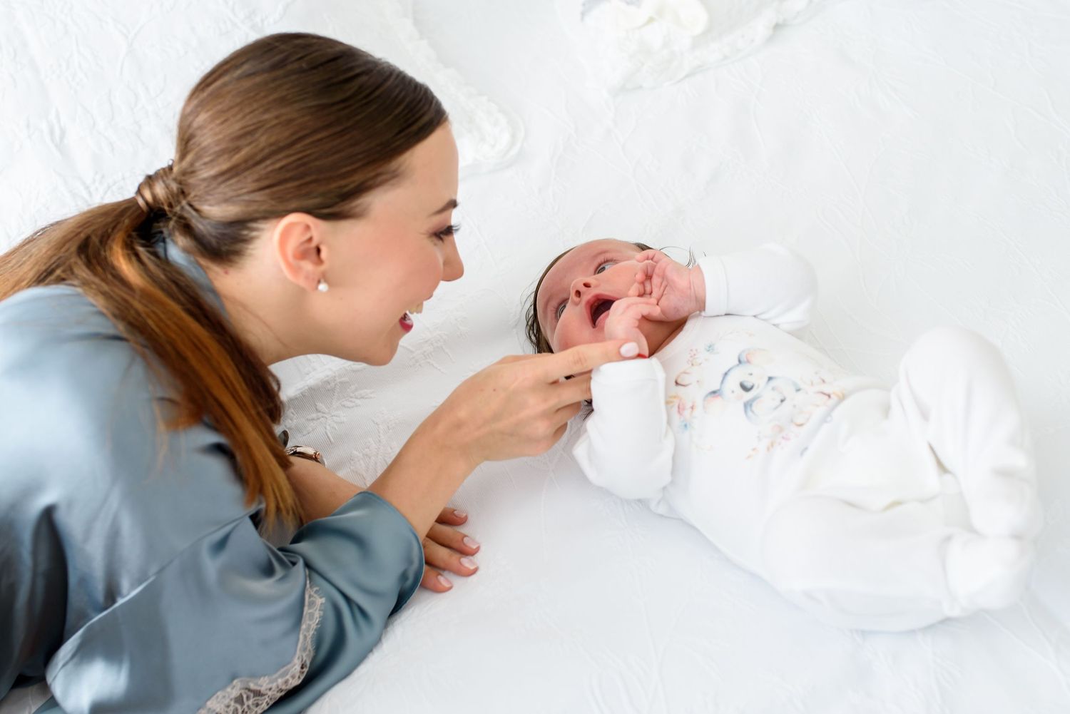 A young mother in a blue satin robe lovingly interacts with her newborn baby lying on a white surface.